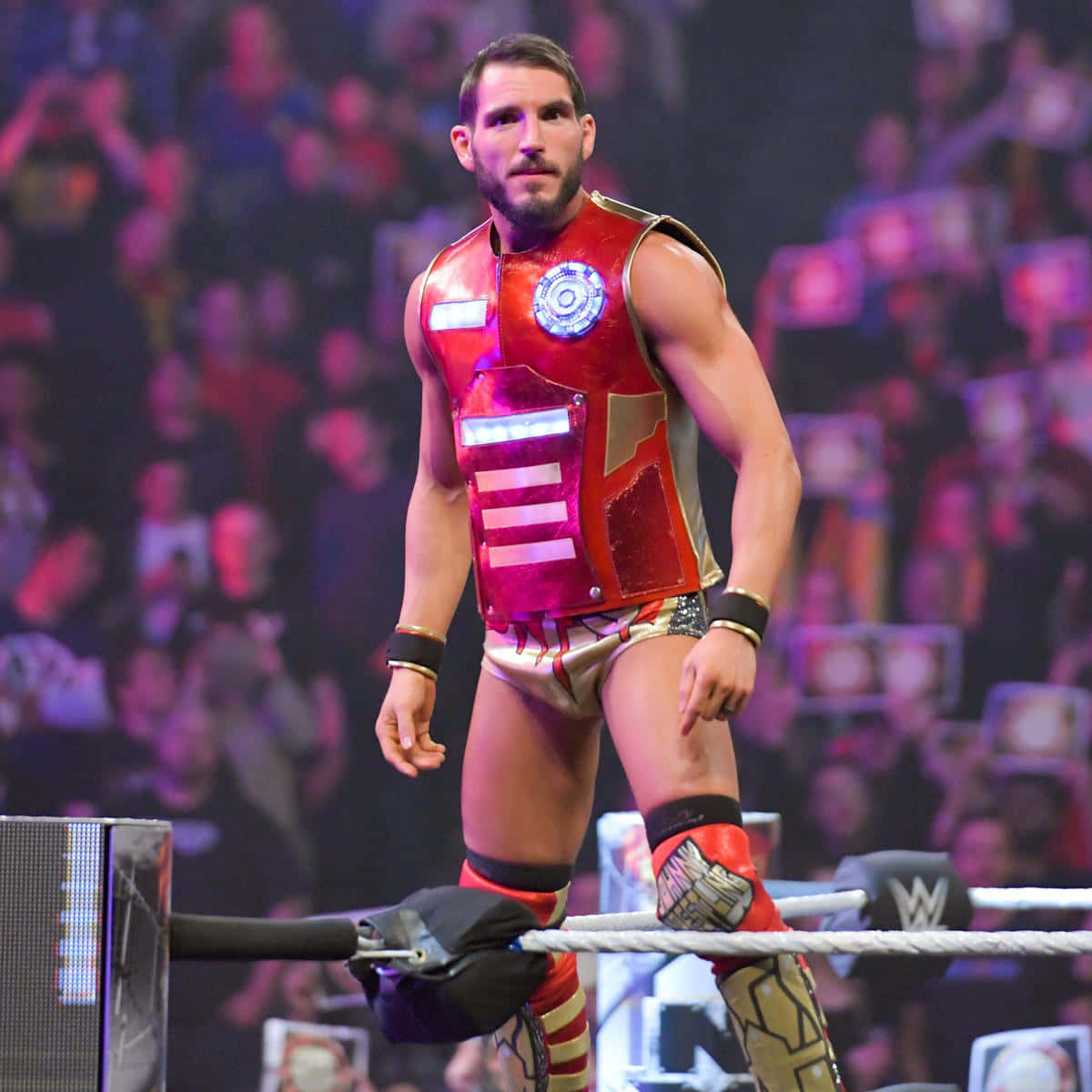 WWE 桌布 壁紙 — 穿著紅色金屬鋼鐵人風格裝備的摔角手自信地站在燈光照射的摔角場中，背後是模糊的人群。