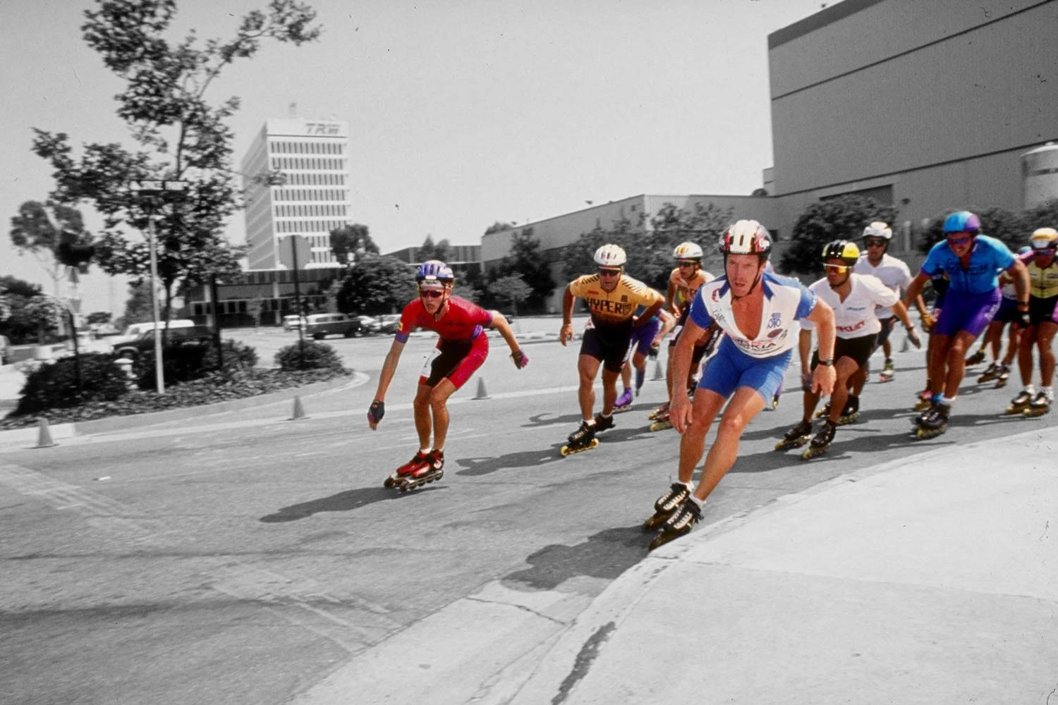 skating 桌布 壁紙 — 穿著色彩繽紛裝備的溜冰者在鋪砌的城市街道上激烈競賽，背景有建築物和樹木。
