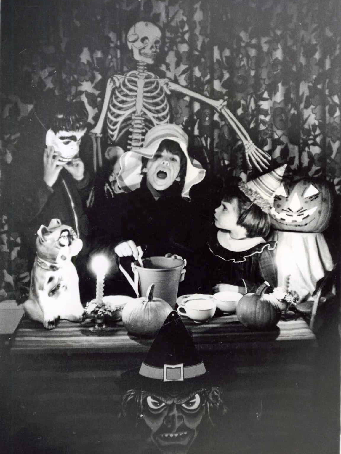 Creepy Halloween 桌布 壁紙 — 穿著萬聖節服裝的孩子們圍繞著一張放有南瓜和蠟燭的桌子，身後有一個骷髏若隱若現。