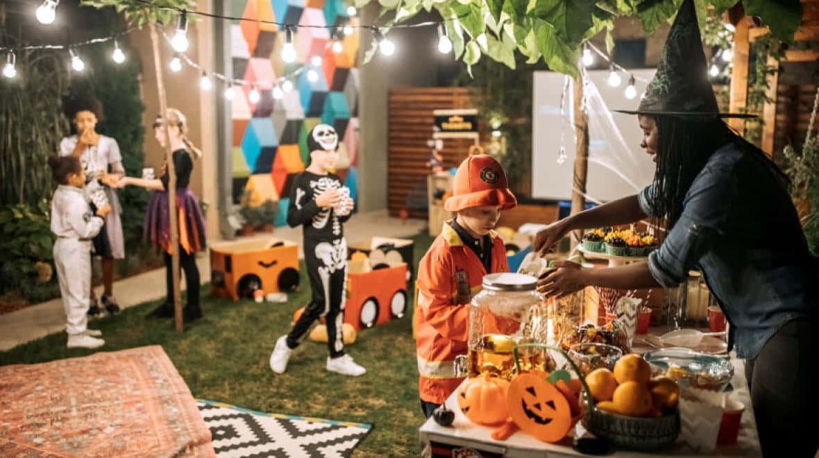 Party 桌布 壁紙 — 穿著萬聖節服裝的孩子們聚集在戶外派對桌旁，上方有燈串和懸掛的樹葉。