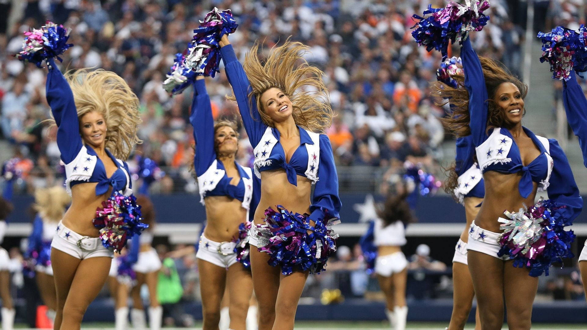 Cheerleader 桌布 壁紙 — 穿著藍白相間、帶有星星裝飾和紫色彩球的啦啦隊員，在模糊的人群背景下的足球場上充滿活力地表演。