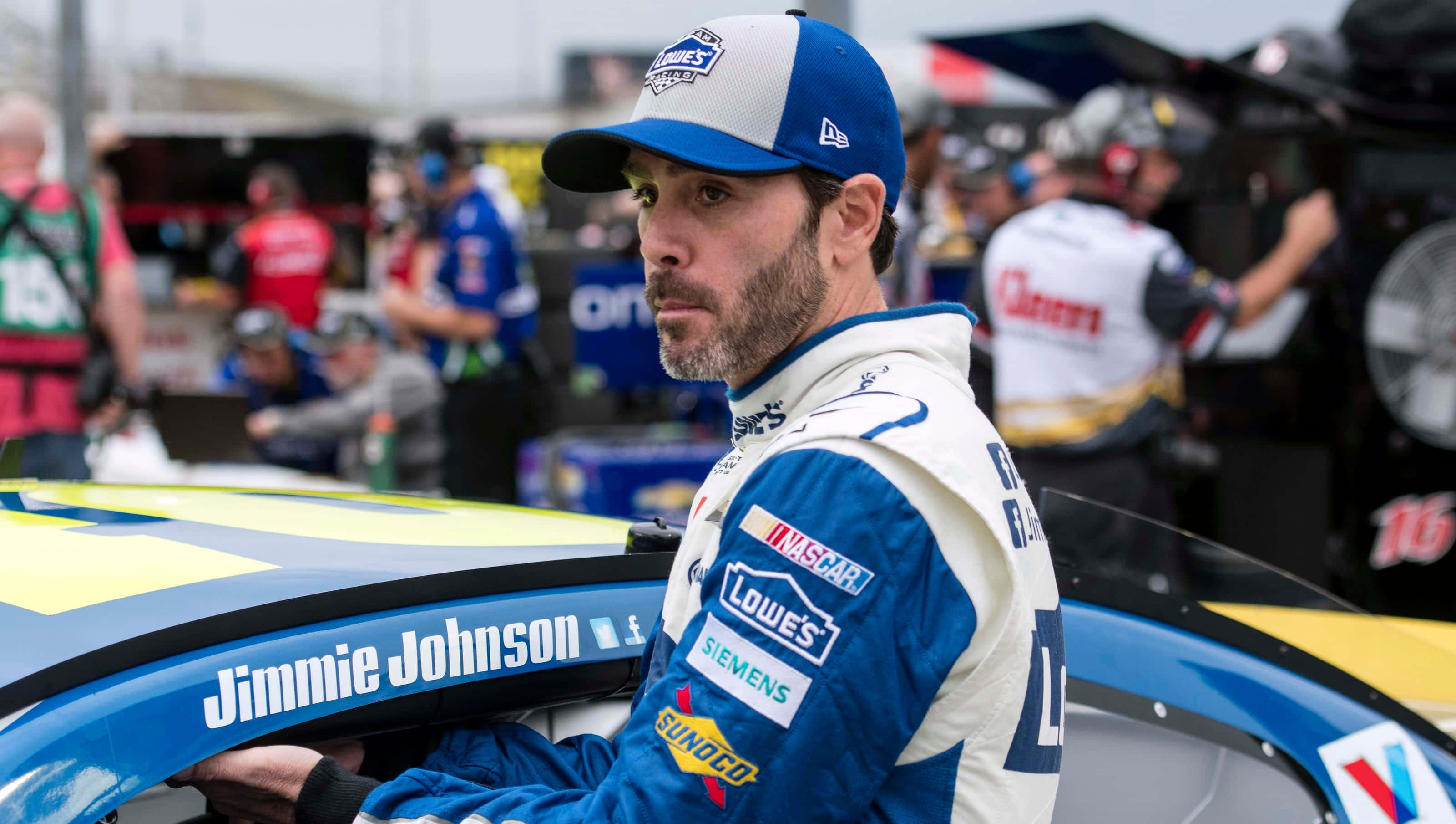 Jimmie Johnson 桌布 壁紙 — 穿著藍白賽車服的Jimmie Johnson在維修站靠在他鮮豔的藍、黃、黑色賽車上。