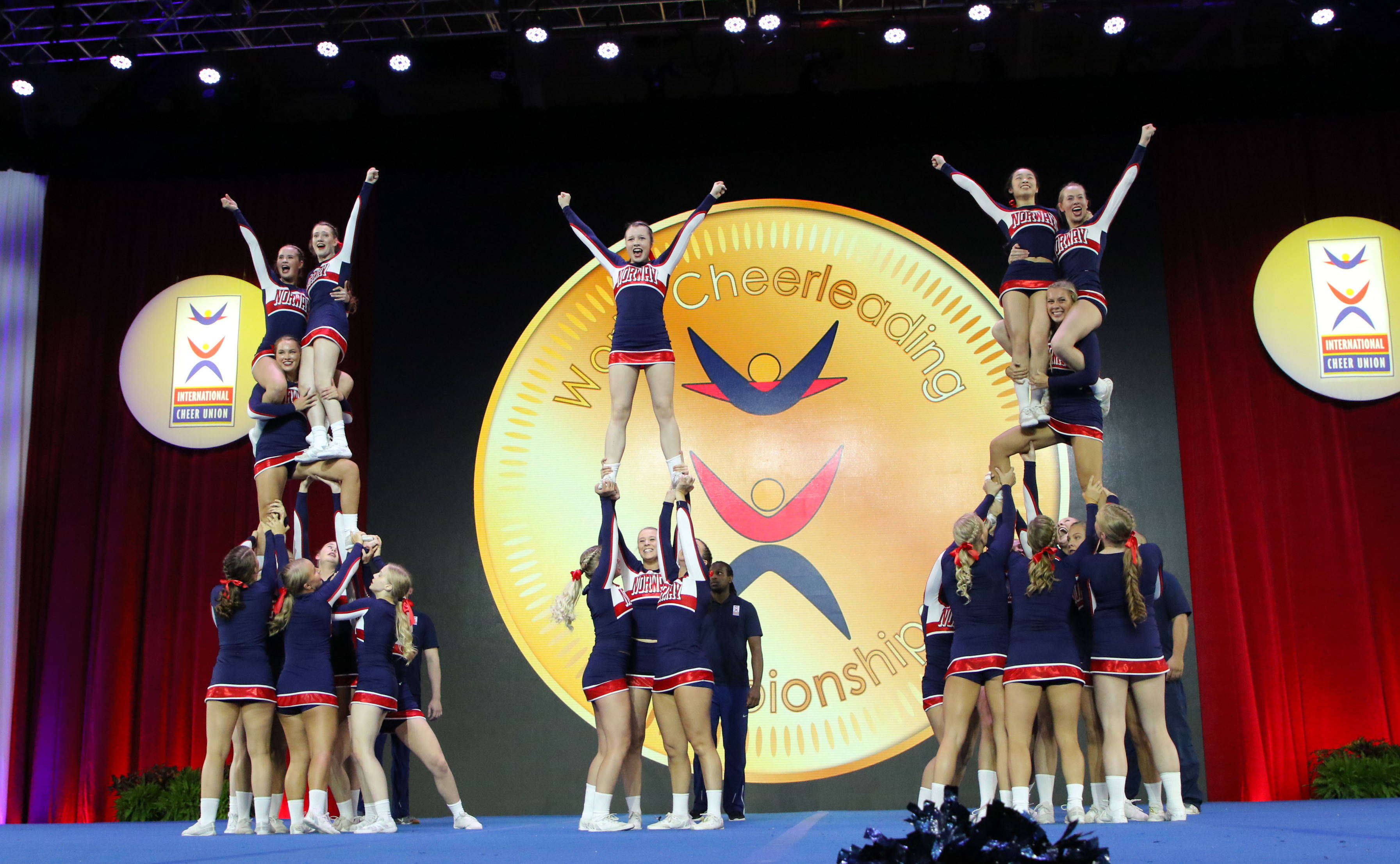 Teamwork 桌布 壁紙 — 穿著藍色和紅色制服的啦啦隊員在一個大型「World Cheerleading Championships」背景前的舞台上形成動態的人體金字塔。