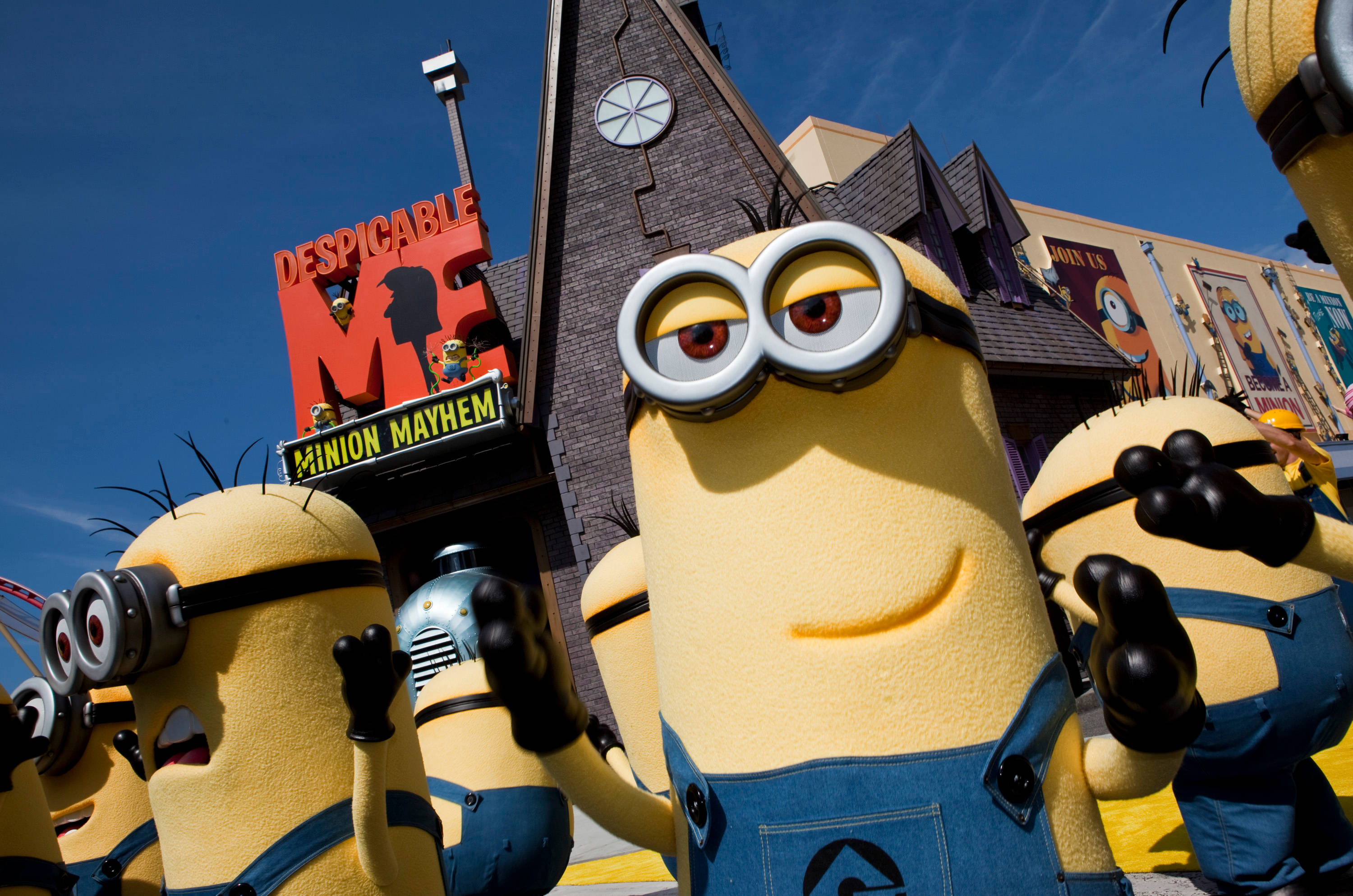 Universal 桌布 壁紙 — 穿著藍色工裝褲的黃色大Minions在晴朗藍天下，於《神偷奶爸》主題建築前開心地揮手。