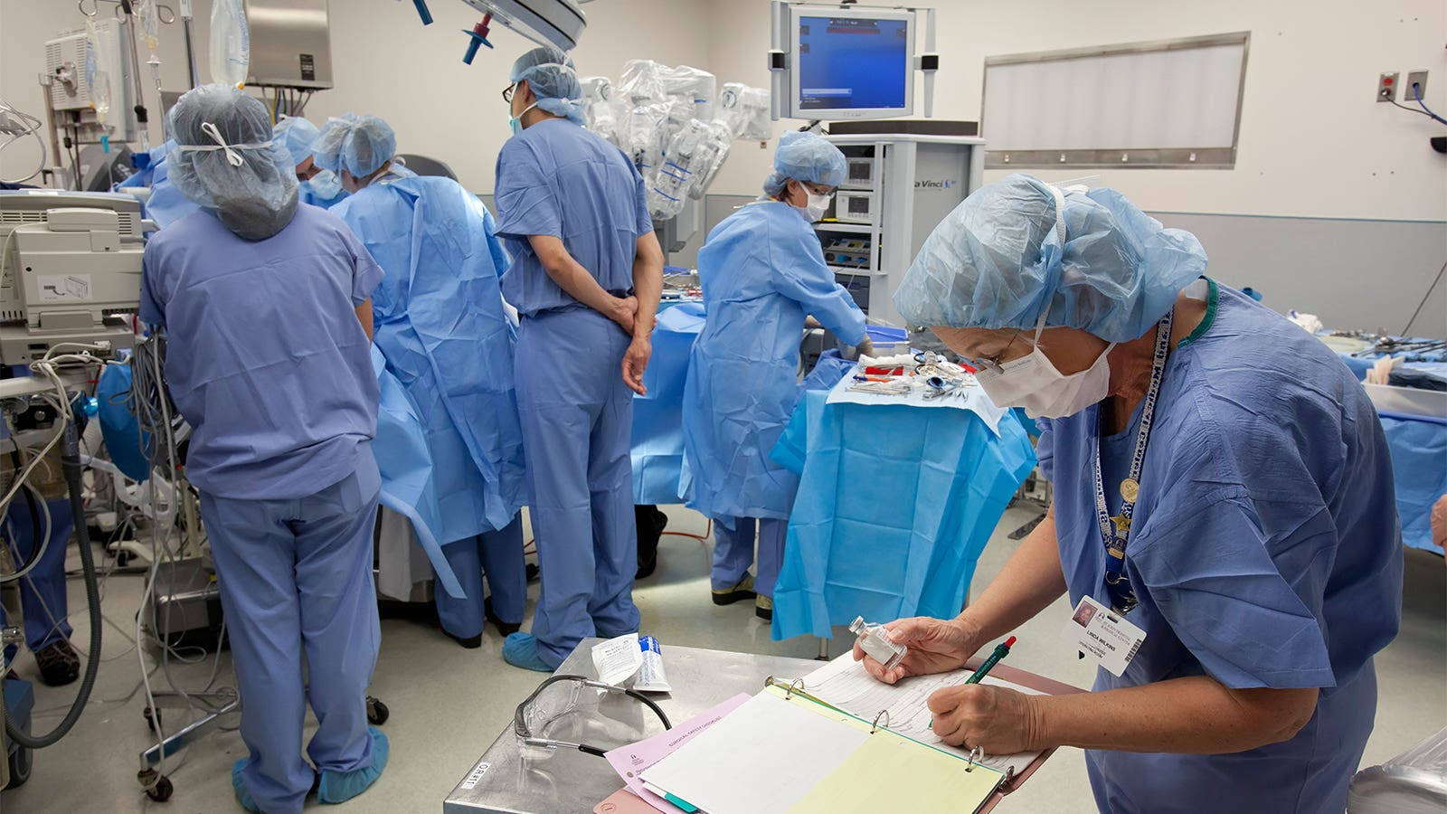 Surgeon 桌布 壁紙 — 穿著藍色手術服和口罩的外科醫生在明亮的、被醫療設備包圍的手術室中專注地工作。