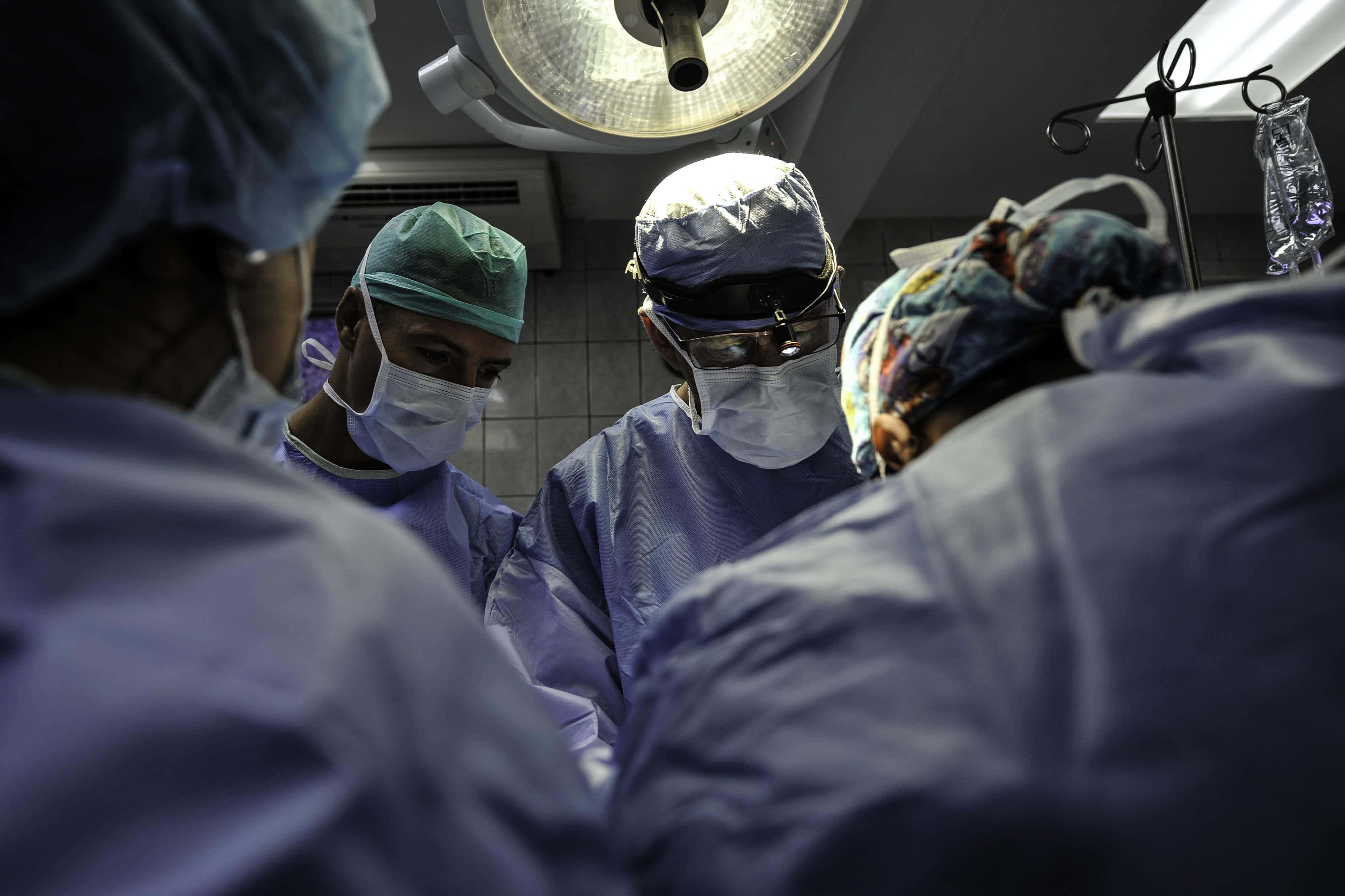 Hospital 桌布 壁紙 — 穿著藍色手術服和口罩的外科醫生在無菌手術室中聚集在明亮的手術燈下。