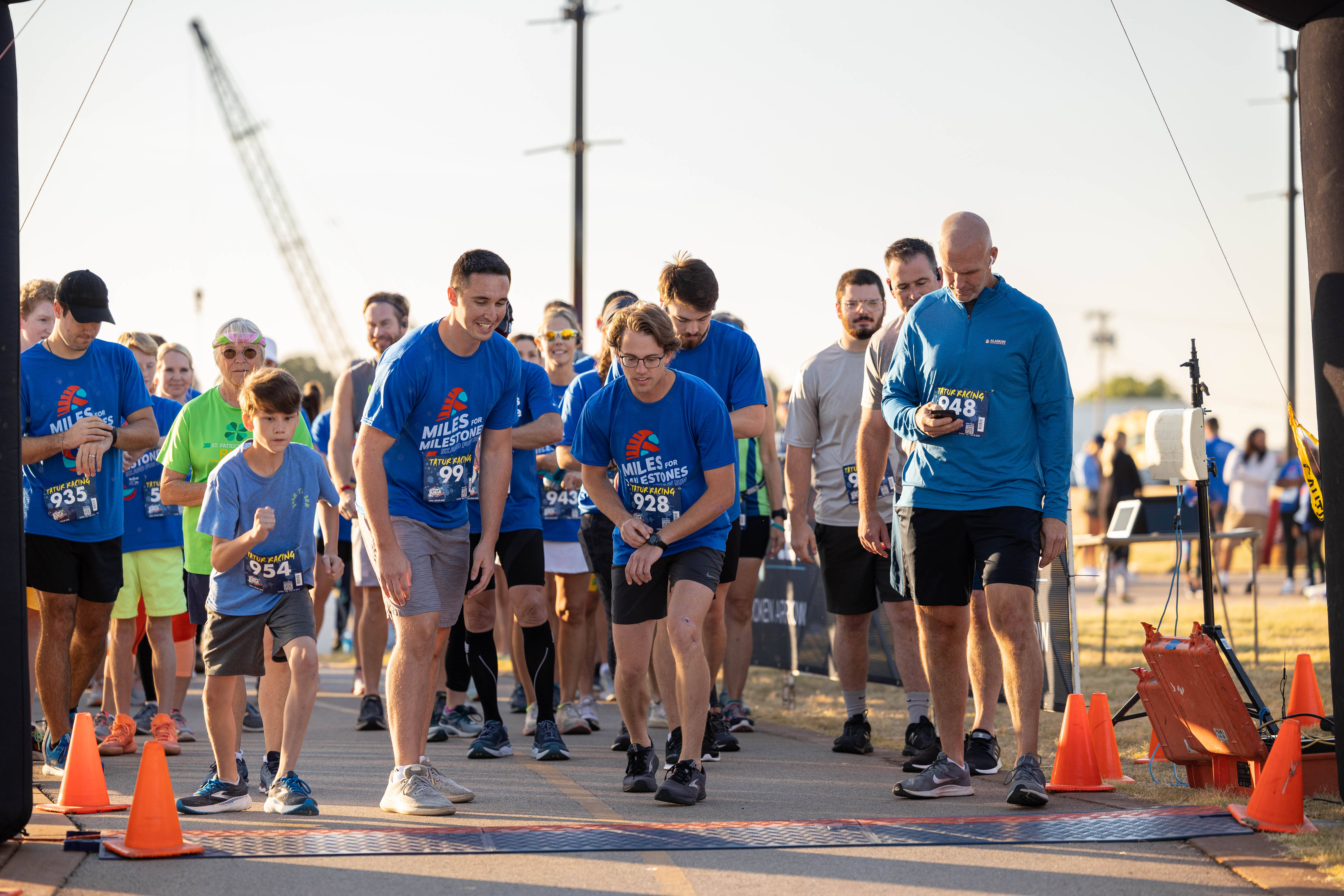 Marathon 桌布 壁紙 — 穿著藍色活動T恤的跑者在明亮的日光下聚集在起跑線，背景有橙色三角錐和起重機。