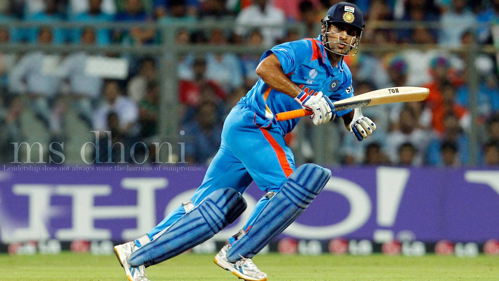 MS Dhoni 桌布 壁紙 — 穿著藍色球衣的板球運動員MS Dhoni在綠色場地上揮動球棒，身後有模糊的人群。