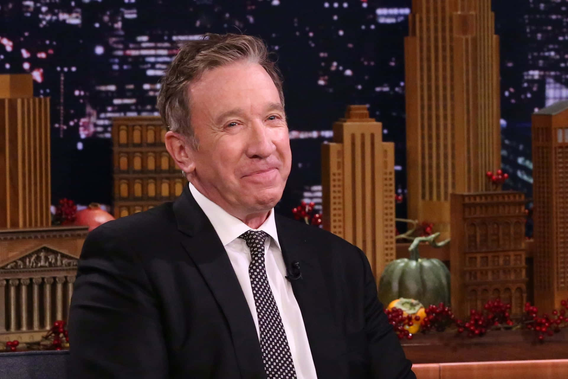 Tim Allen 桌布 壁紙 — 穿著西裝、面露疲態的男子在城市天際線和秋季裝飾的脫口秀場景中微微微笑。