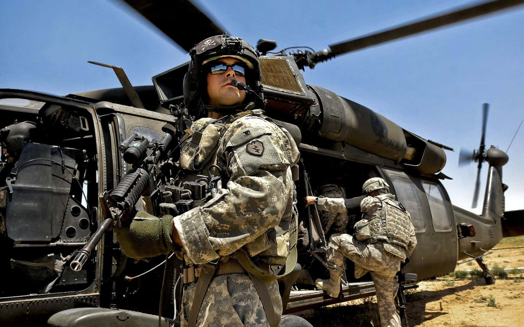 軍隊 桌布 壁紙 — 穿著迷彩裝備的士兵在沙漠環境中，在晴朗的藍天下，警覺地站在一架黑色軍用直升機旁邊。