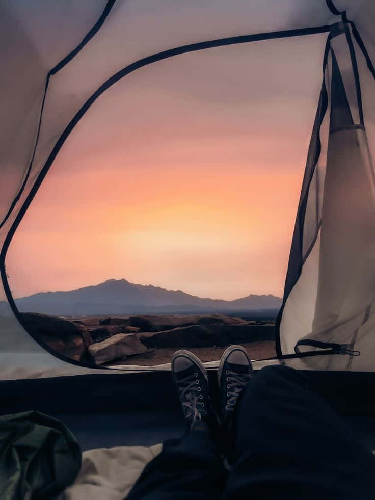 Minimalist Camping 桌布 壁紙 — 穿著運動鞋的雙腳在帳篷內休息，溫暖的夕陽將遠山染上柔和的橙色和粉紅色調。