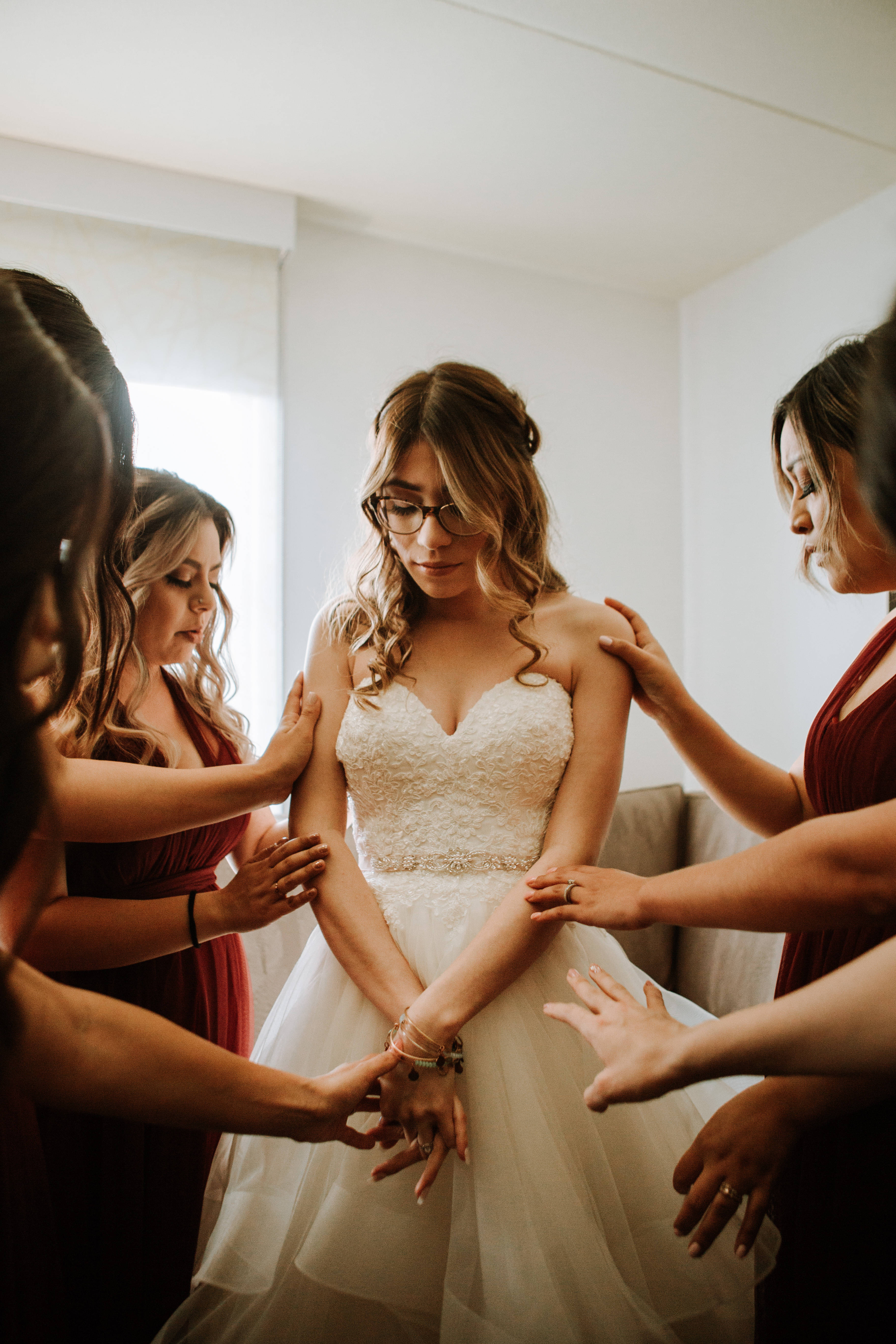 Bridesmaids 桌布 壁紙 — 穿著酒紅色連衣裙的伴娘輕輕地將手放在一位穿著白色蕾絲禮服的新娘身上，她站在一個光線柔和的房間裡。