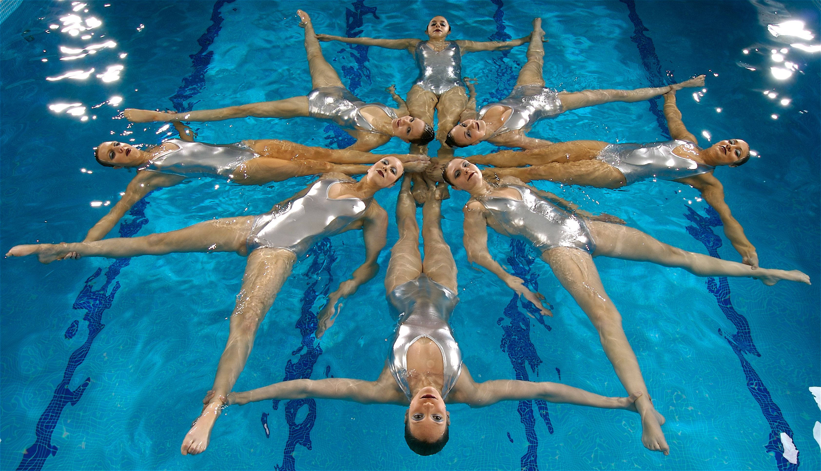 Swimming 桌布 壁紙 — 穿著銀色泳衣的韻律泳選手在充滿活力的藍色泳池表面形成一個閃爍的星爆圖案。