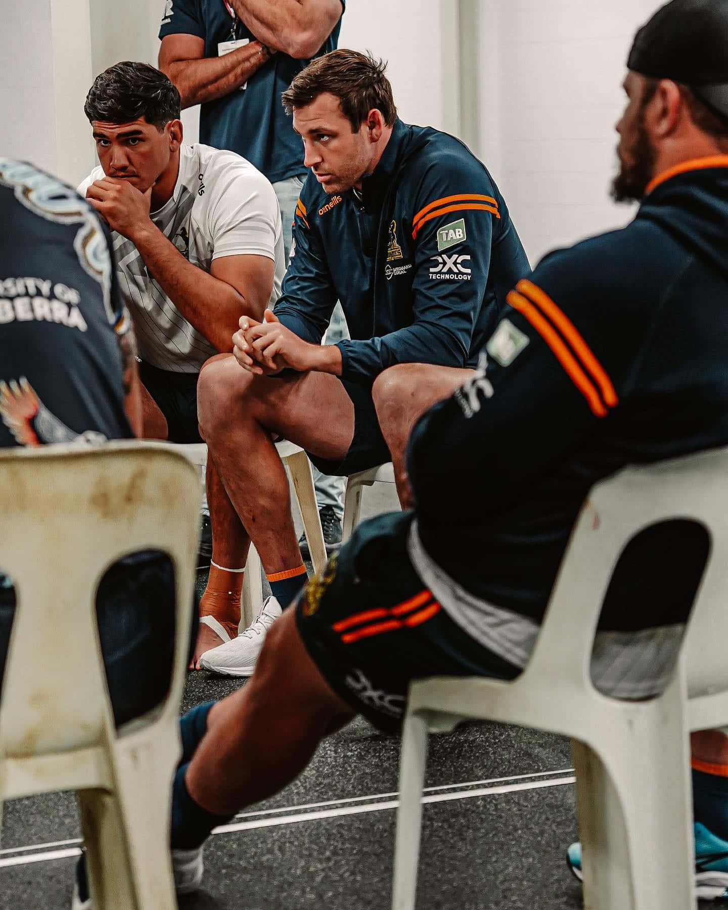 Brumbies 桌布 壁紙 — 穿著隊服的運動員坐在更衣室的塑膠椅上，專注而嚴肅地準備比賽。