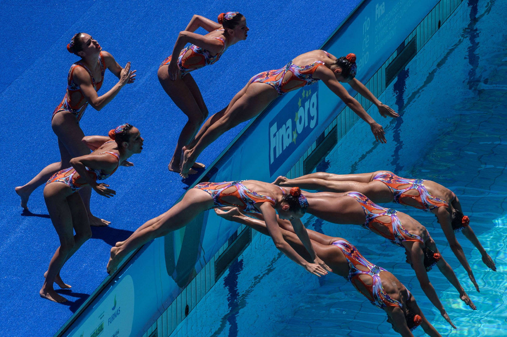 Swimming 桌布 壁紙 — 穿著鮮豔橙色和藍色服裝的韻律泳選手從泳池邊一躍而起，在明亮的藍色水面上形成一道動態弧線。
