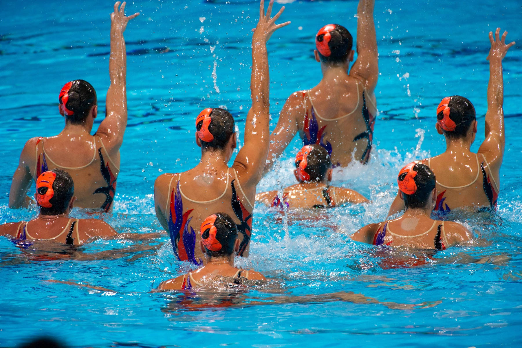 Swimming 桌布 壁紙 — 穿著鮮豔泳衣的韻律泳選手一致舉起手臂，在明亮的藍色泳池水中營造出動態水花。