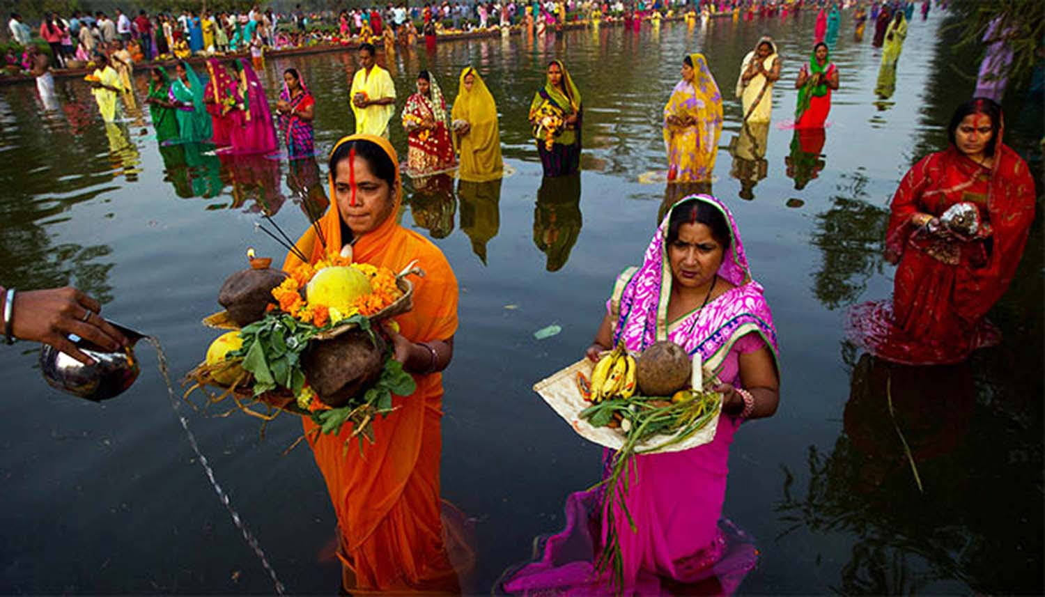Indian 桌布 壁紙 — 穿著鮮豔紗麗的女性在文化儀式中涉水過河，手持鮮花、水果和椰子的祭品。