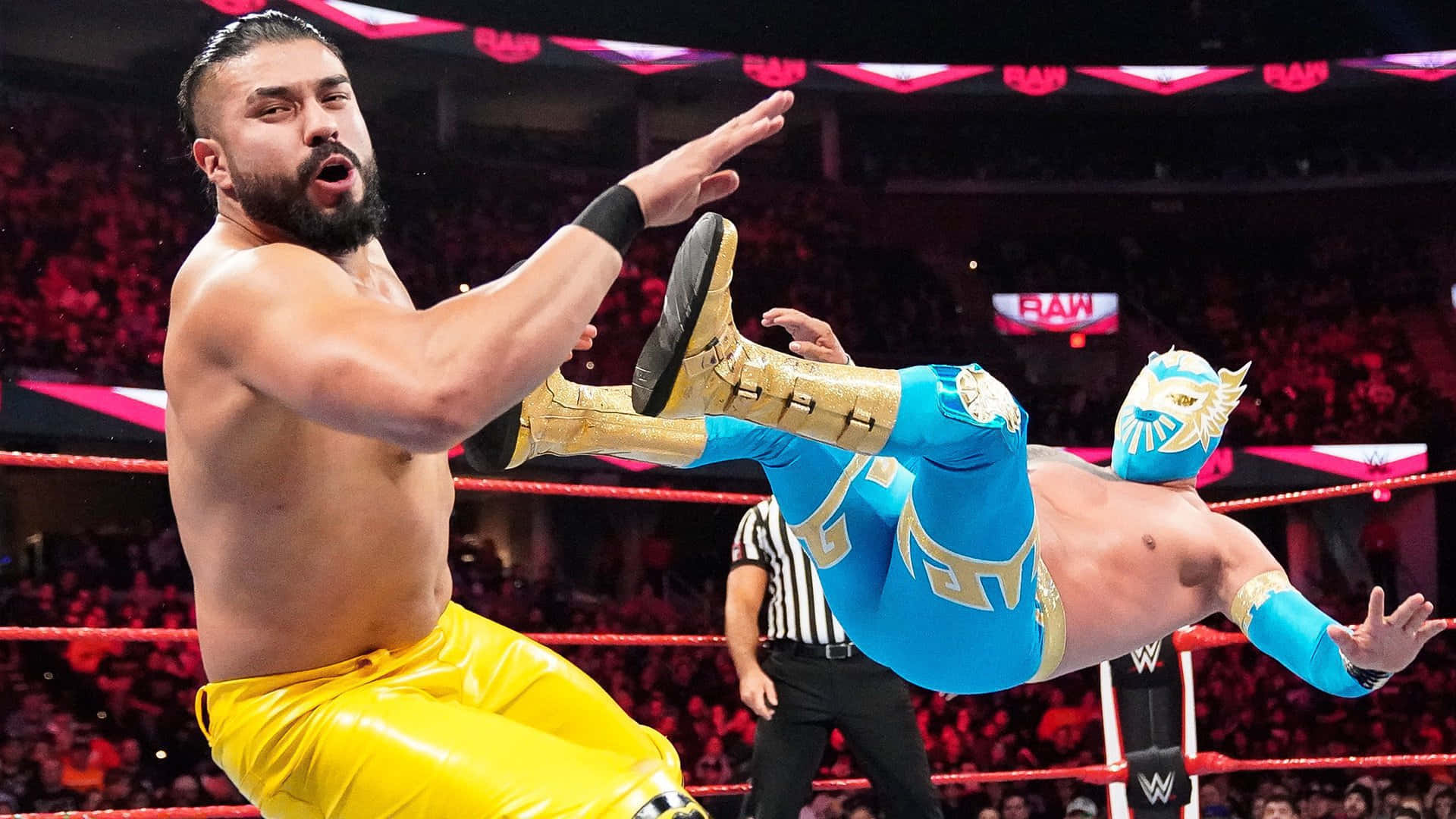 WWE 桌布 壁紙 — 穿著黃色緊身衣的摔角手在燈光明亮的摔角場內對身穿藍金色裝備的對手執行高空飛行動作。