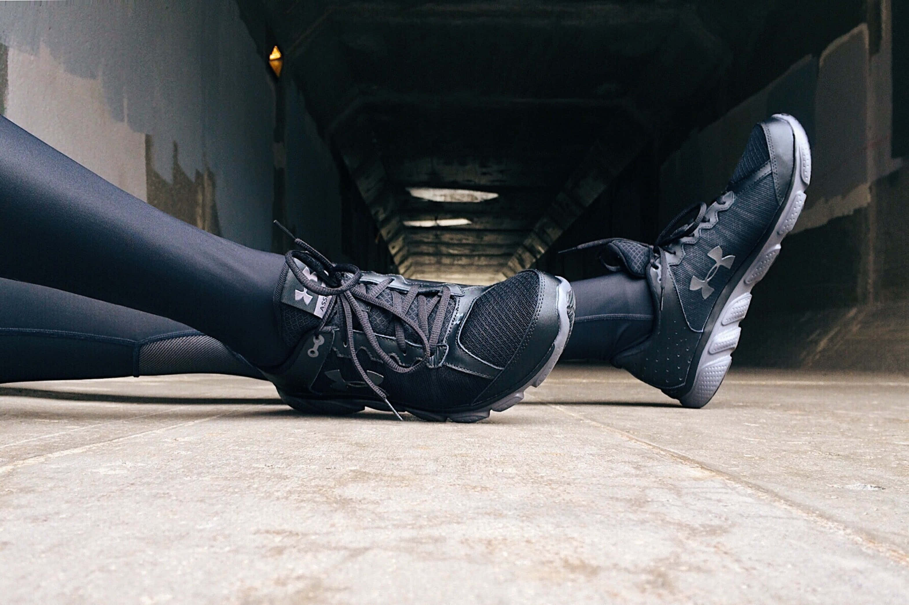 Under Armour 桌布 壁紙 — 穿著黑色Under Armour運動鞋的運動員腿部特寫，在昏暗的城市隧道下休息在混凝土上。