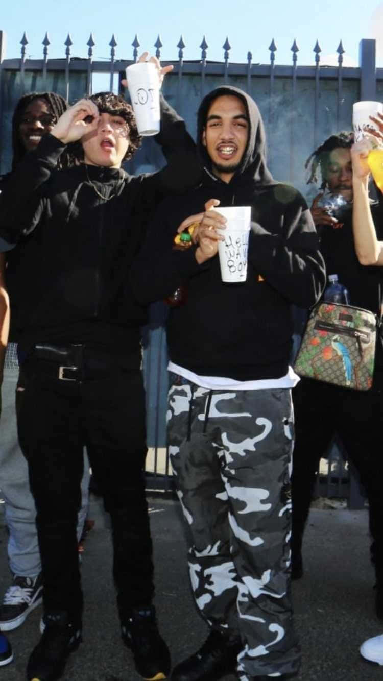 Shoreline Mafia 桌布 壁紙 — 穿著黑色連帽衫和迷彩褲的年輕人，在明亮的日光下，對著帶刺的金屬圍欄愉快地舉杯慶祝。