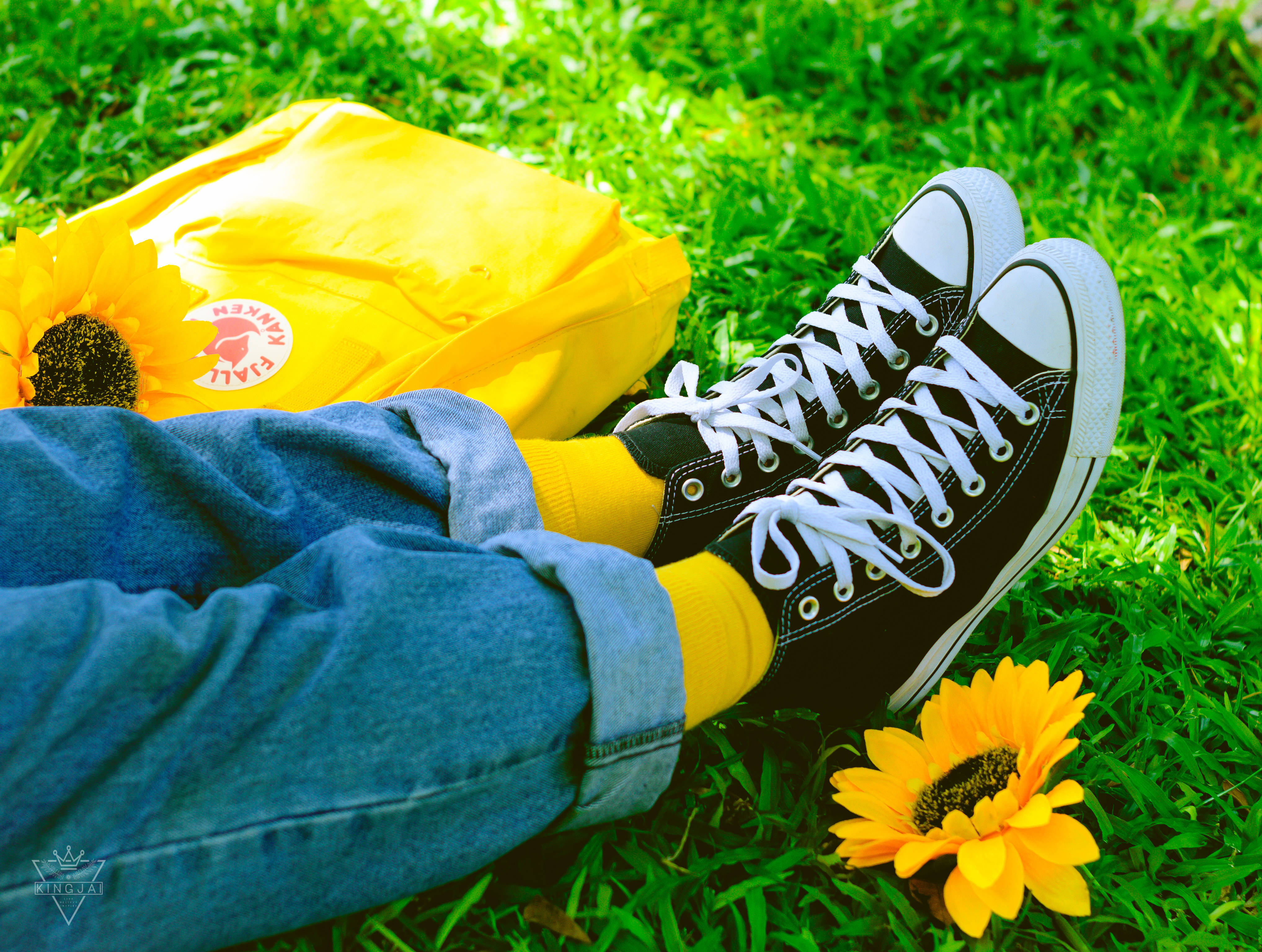 Converse 桌布 壁紙 — 穿著黑色運動鞋和黃色襪子的放鬆人物在充滿活力的綠色草地上休息，旁邊有一個黃色背包和向日葵。