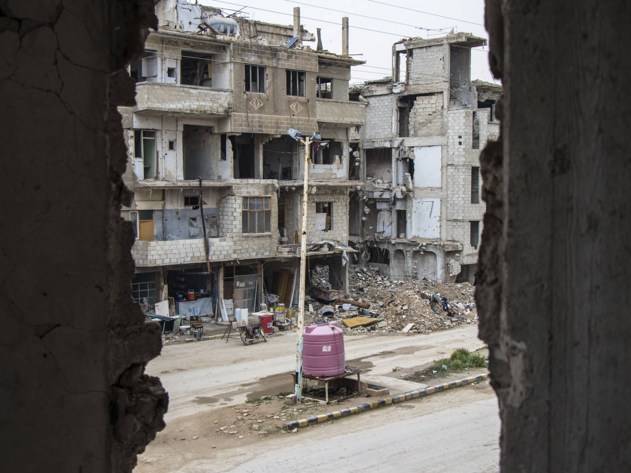 Syria 桌布 壁紙 — 穿過破損的牆框，一條荒涼的街道在陰天之下顯露出廢棄的多層建築和瓦礫。