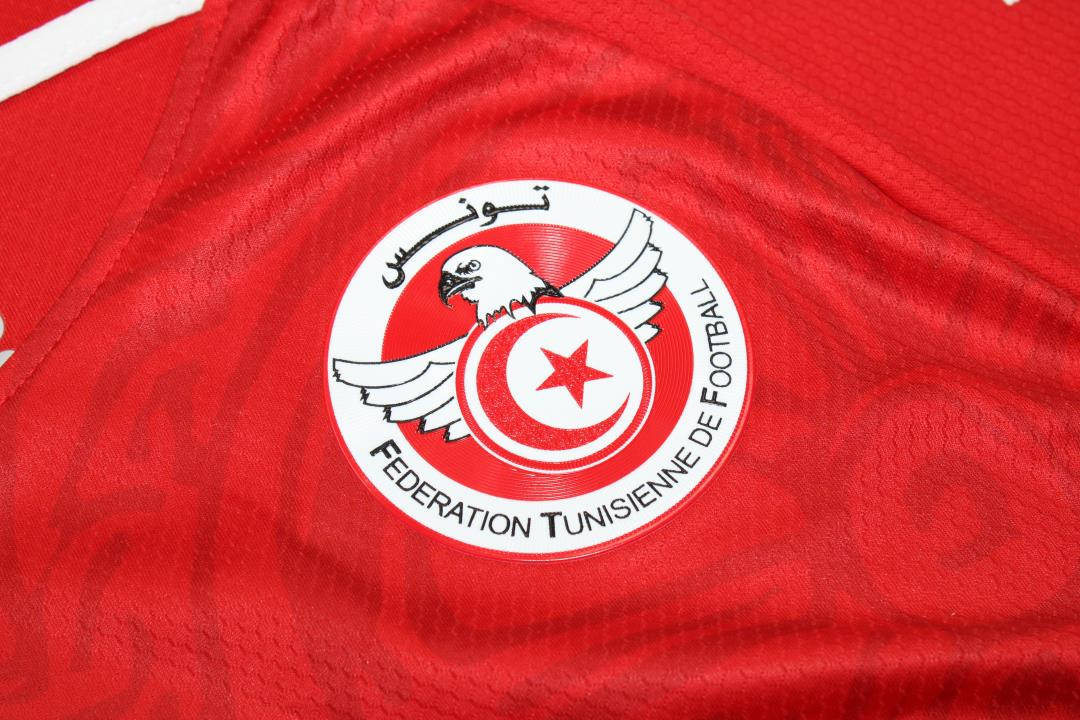 Tunisia 桌布 壁紙 — 突尼西亞足球協會徽章在帶有紋理的紅色球衣布料上的特寫，可見縫線和皺褶。