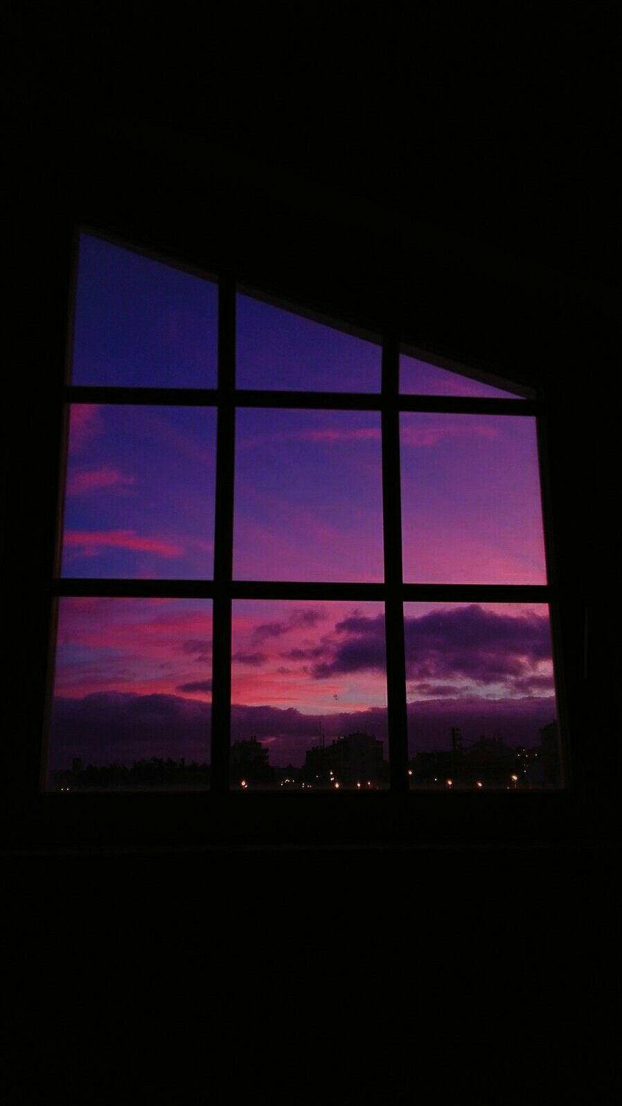 Clouds Aesthetics 桌布 壁紙 — 窗戶框住了一片在暮色中遠處城市燈光上方的 vibrant 紫色和粉紅色日落天空。