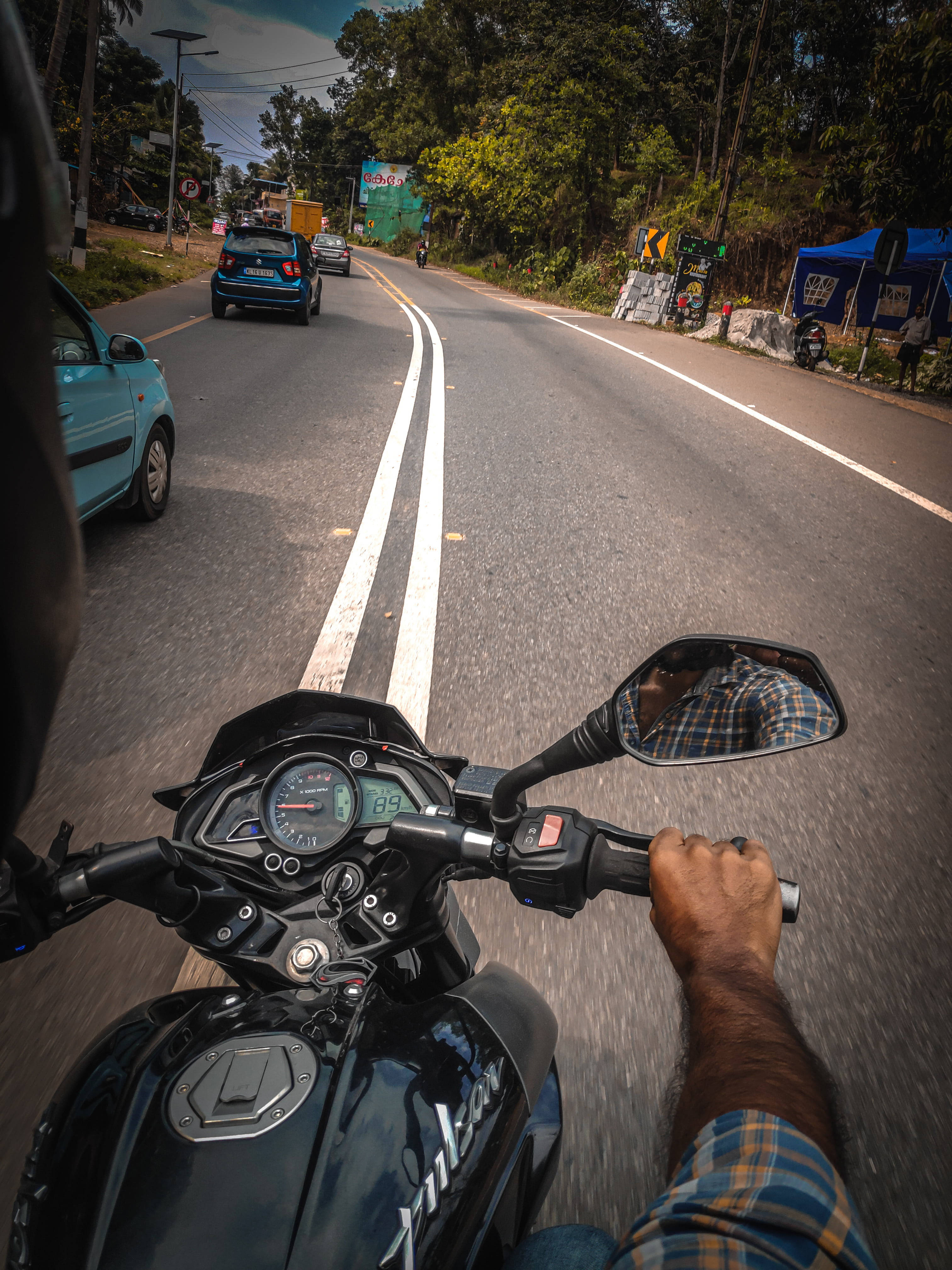 Road 桌布 壁紙 — 第一人稱視角，一名騎士握著黑色摩托車的把手，在蜿蜒的柏油路上疾馳，路邊有樹木和攤位。
