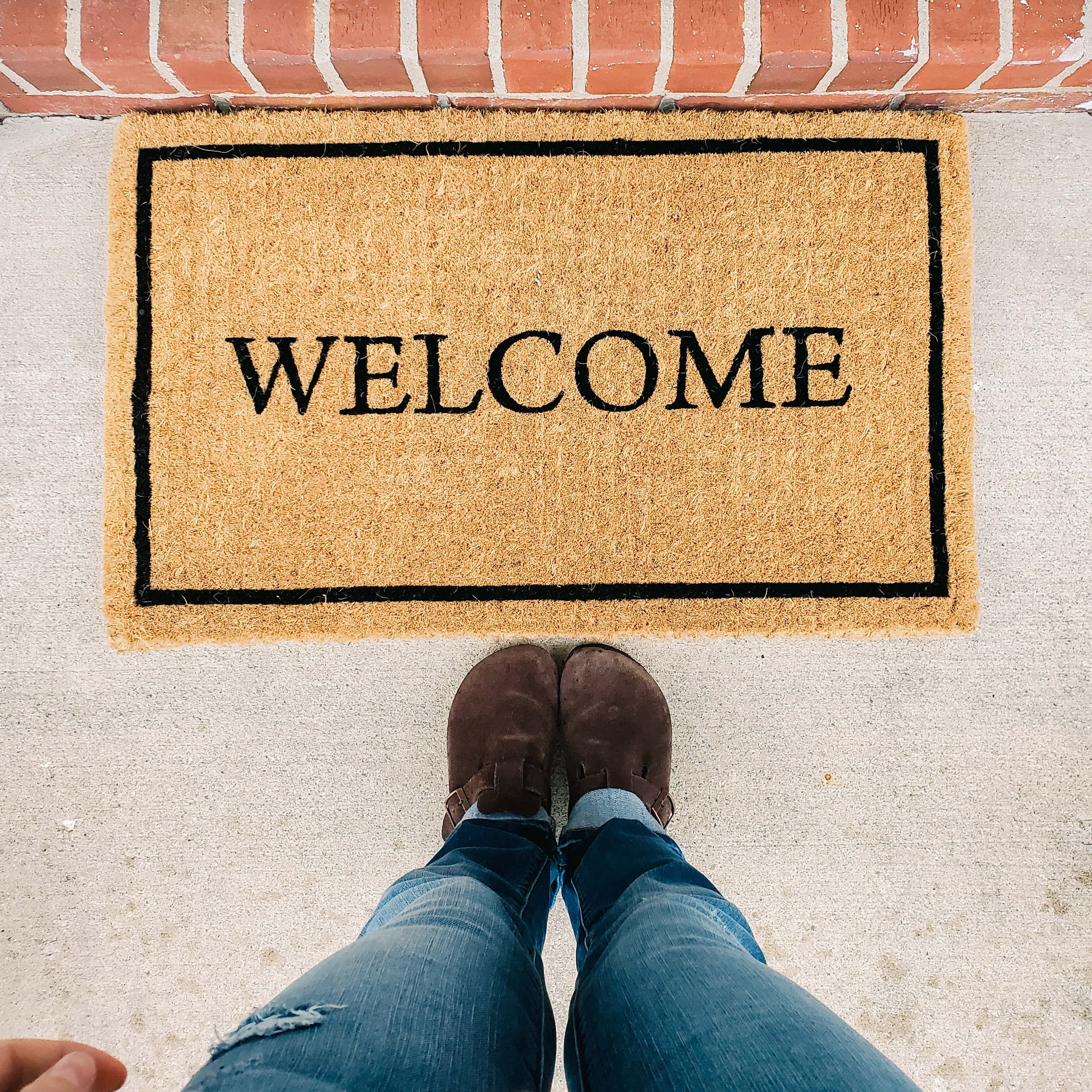 Welcome 桌布 壁紙 — 第一人稱視角，棕色鞋子踏在紅磚門階前的棕褐色迎賓地墊上。