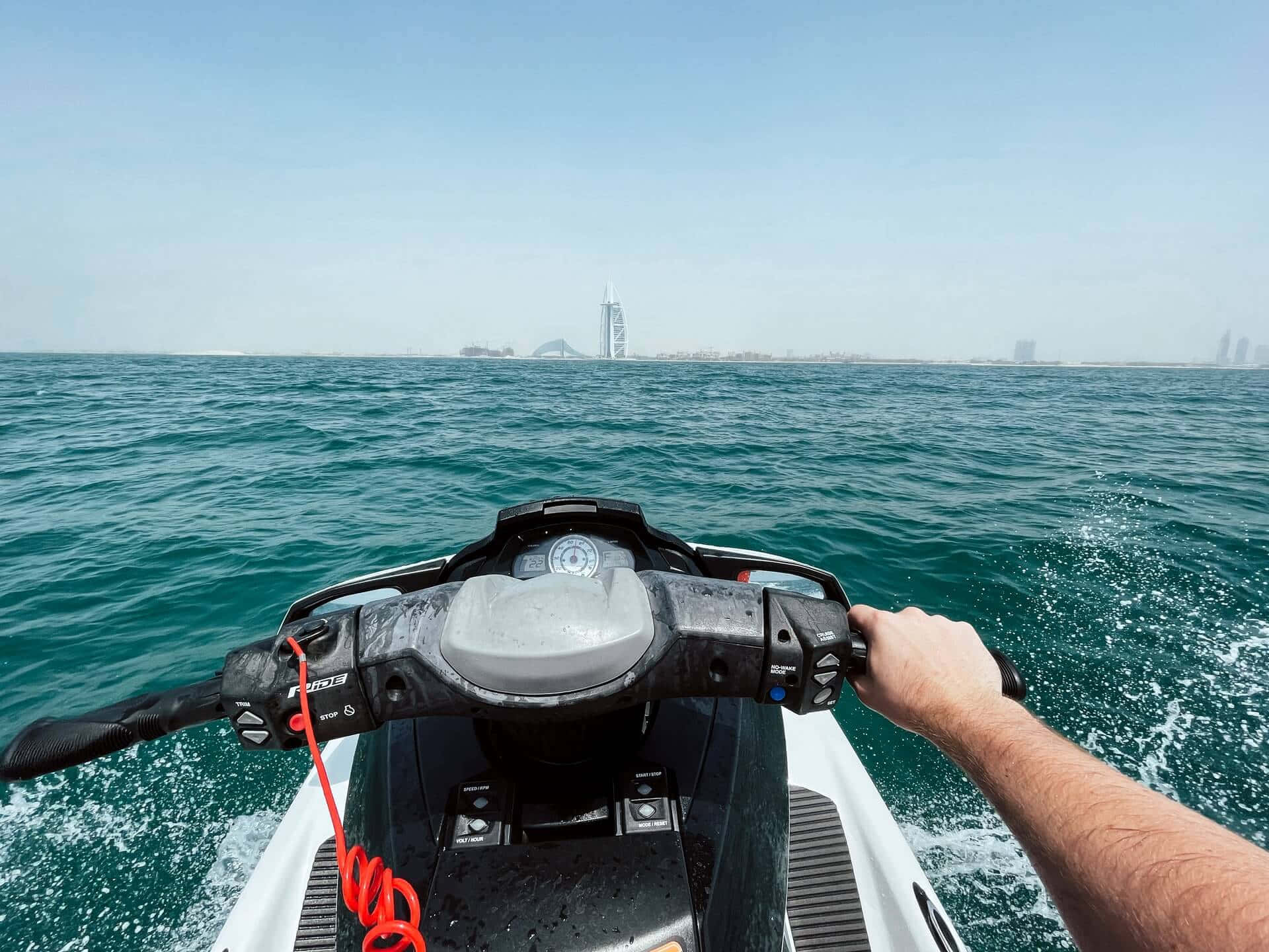 adventure 桌布 壁紙 — 第一人稱視角，駕駛水上摩托車穿越碧綠海水，朝向杜拜標誌性的帆船飯店天際線，天空晴朗湛藍。