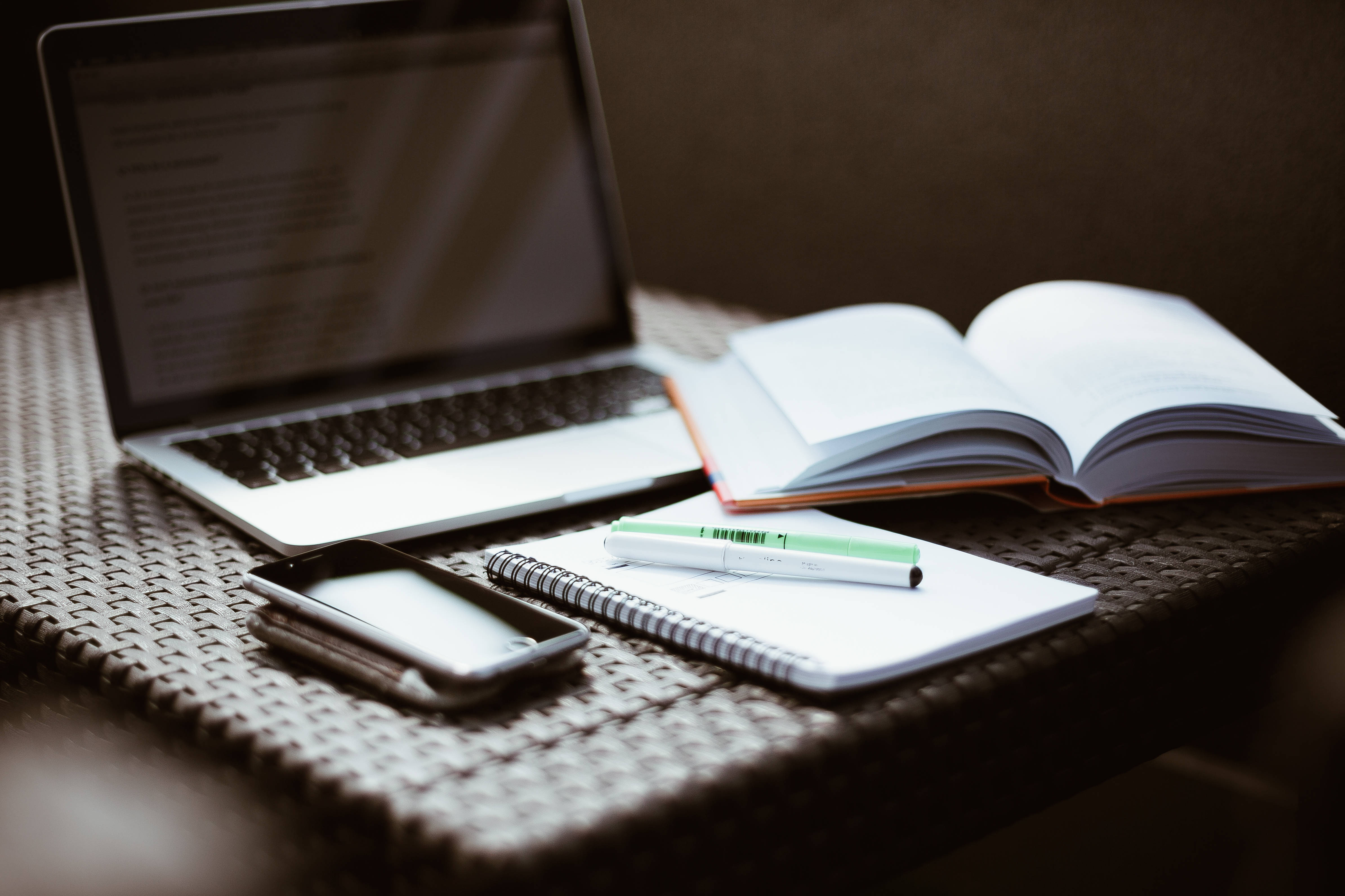 Studying 桌布 壁紙 — 筆記型電腦、筆記本和電話放在編織桌上，旁邊有一本打開的書，在溫暖的環境光下。