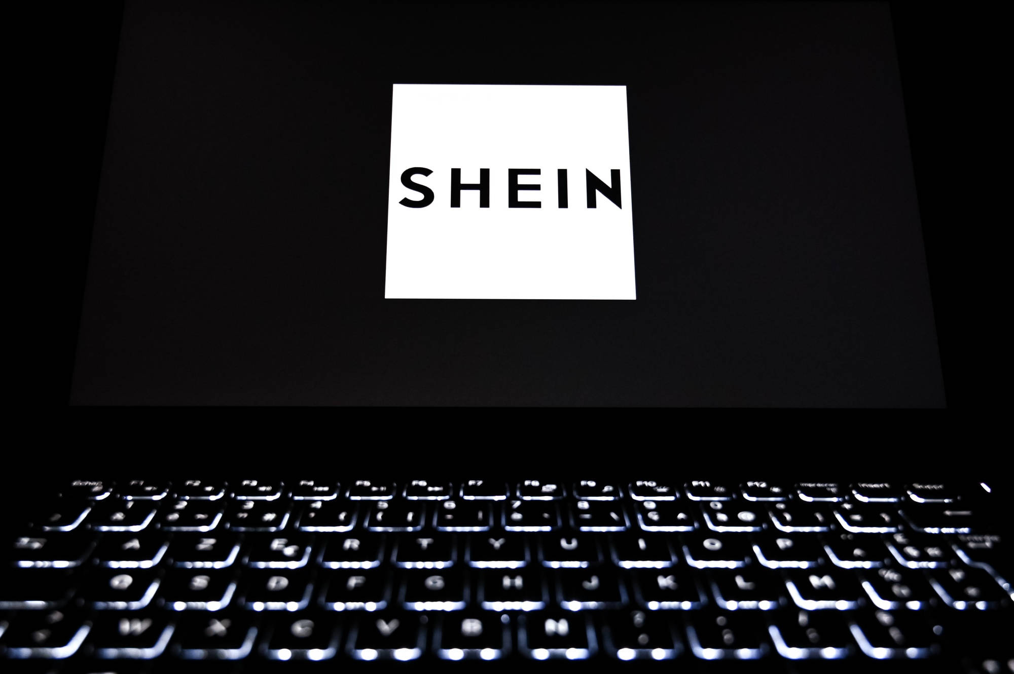 Shein 桌布 壁紙 — 筆記型電腦鍵盤在黑暗中發出微弱光芒，螢幕上顯示黑色背景上鮮明的白色SHEIN標誌。