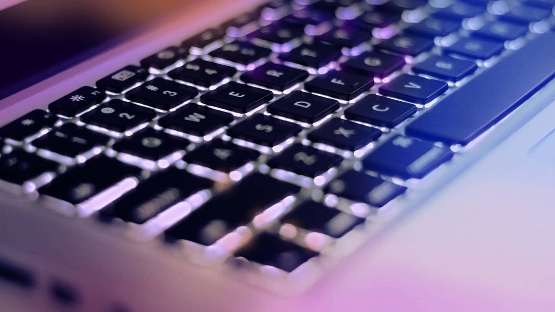 美學 MacBook 壁紙 桌布 壁紙 — 筆記型電腦鍵盤的特寫，沐浴在柔和的紫色和藍色環境光中，景深較淺。
