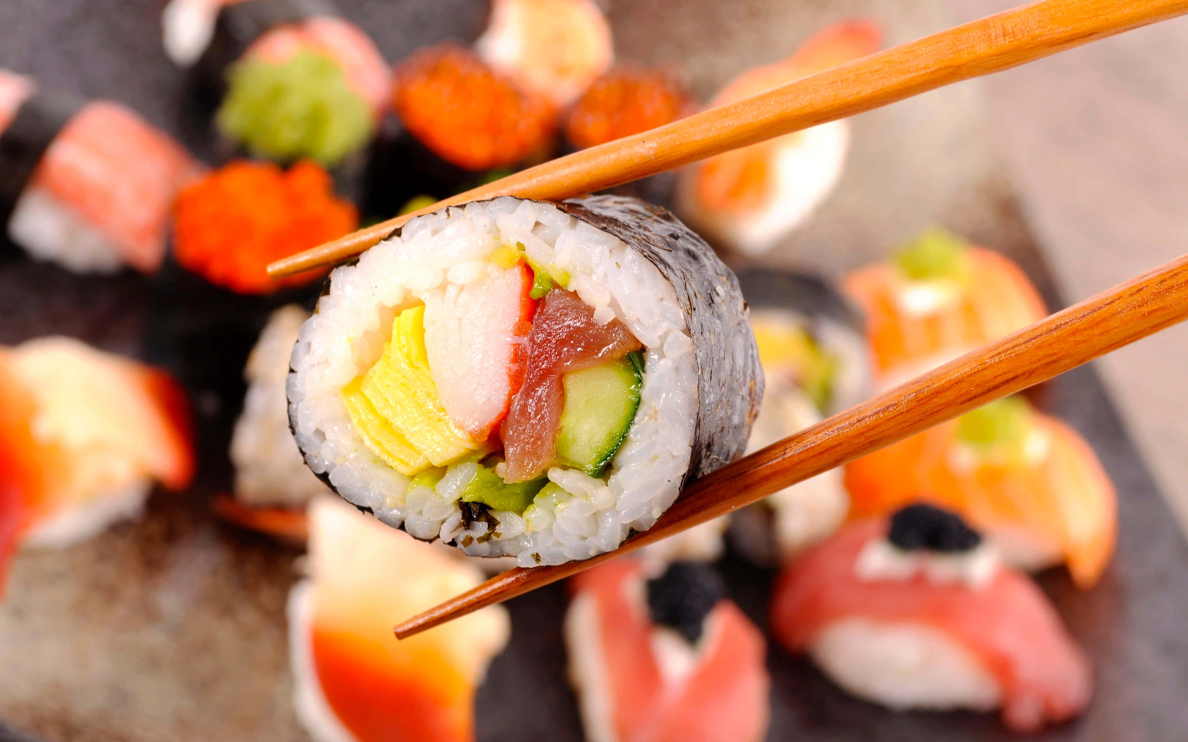 Japanese Aesthetic HD 桌布 壁紙 — 筷子夾住壽司卷（內含米飯、鮪魚、小黃瓜和雞蛋）的特寫，背景是模糊的各式壽司。