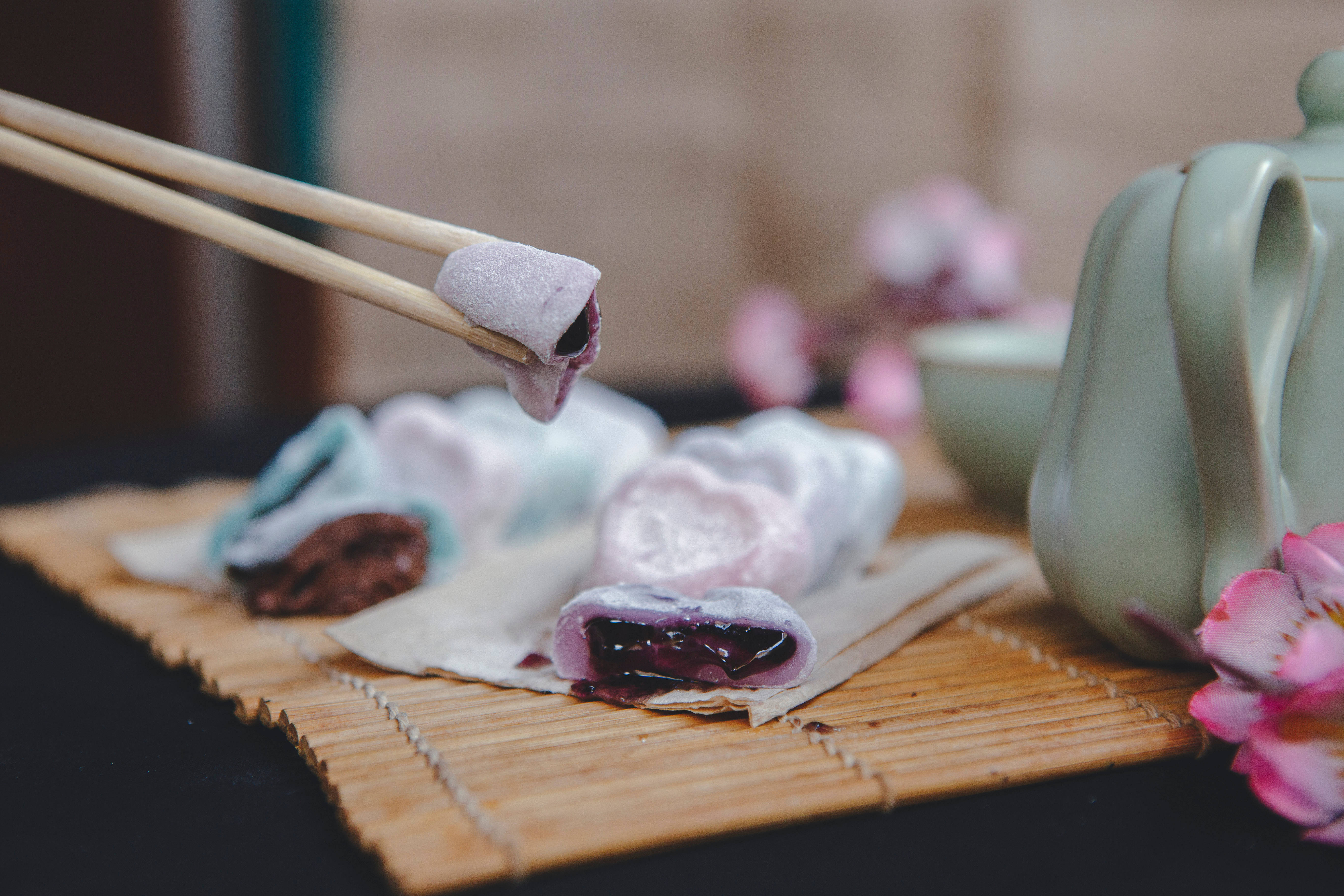 Mochi 桌布 壁紙 — 筷子夾起一個紫色麻糬糰子，露出深色莓果餡料，旁邊有綠色茶壺和粉紅櫻花在竹蓆上。