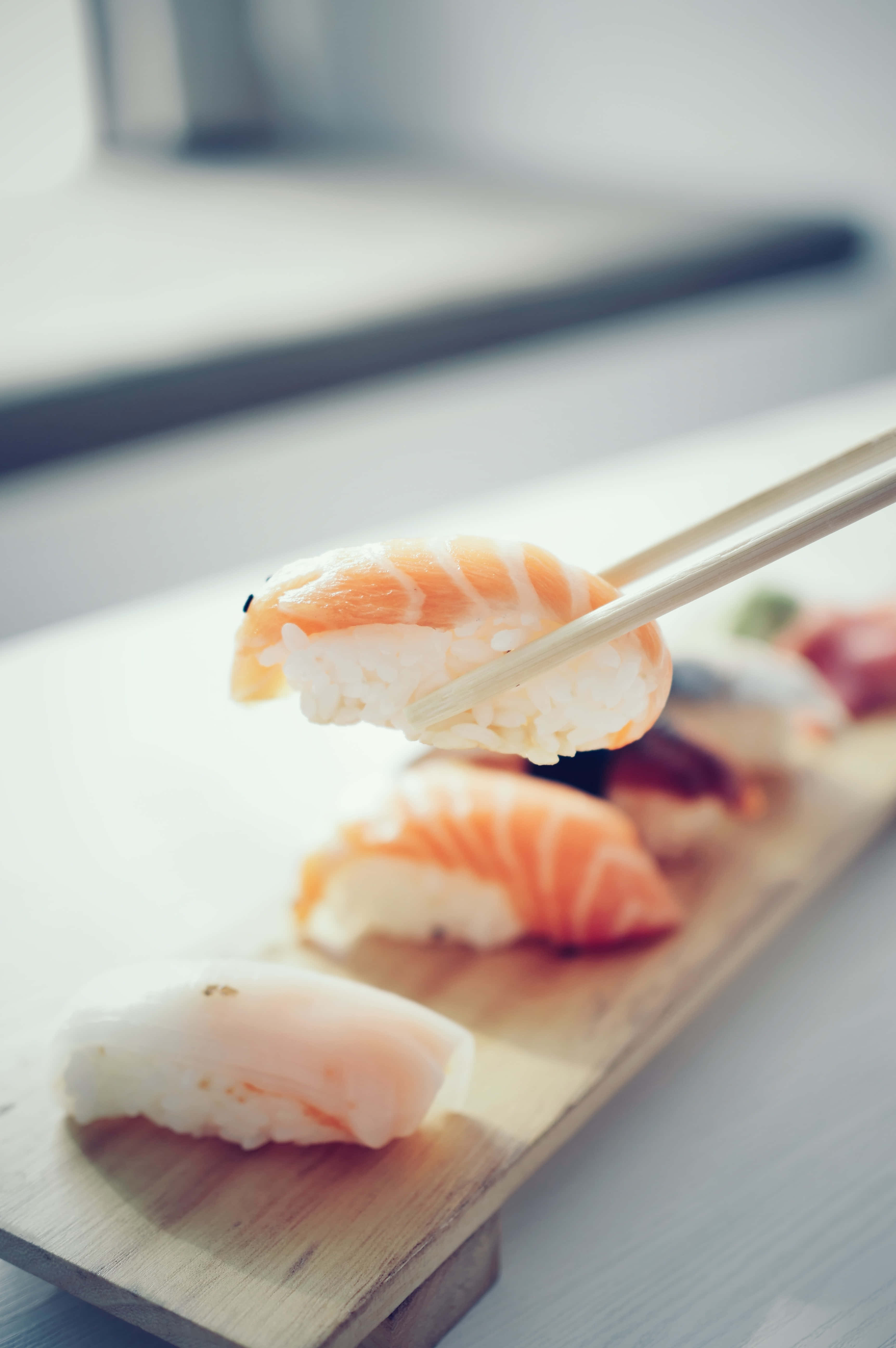 Sushi 桌布 壁紙 — 筷子從木盤上夾起一片鮭魚握壽司的特寫，背景中其他壽司片模糊不清。