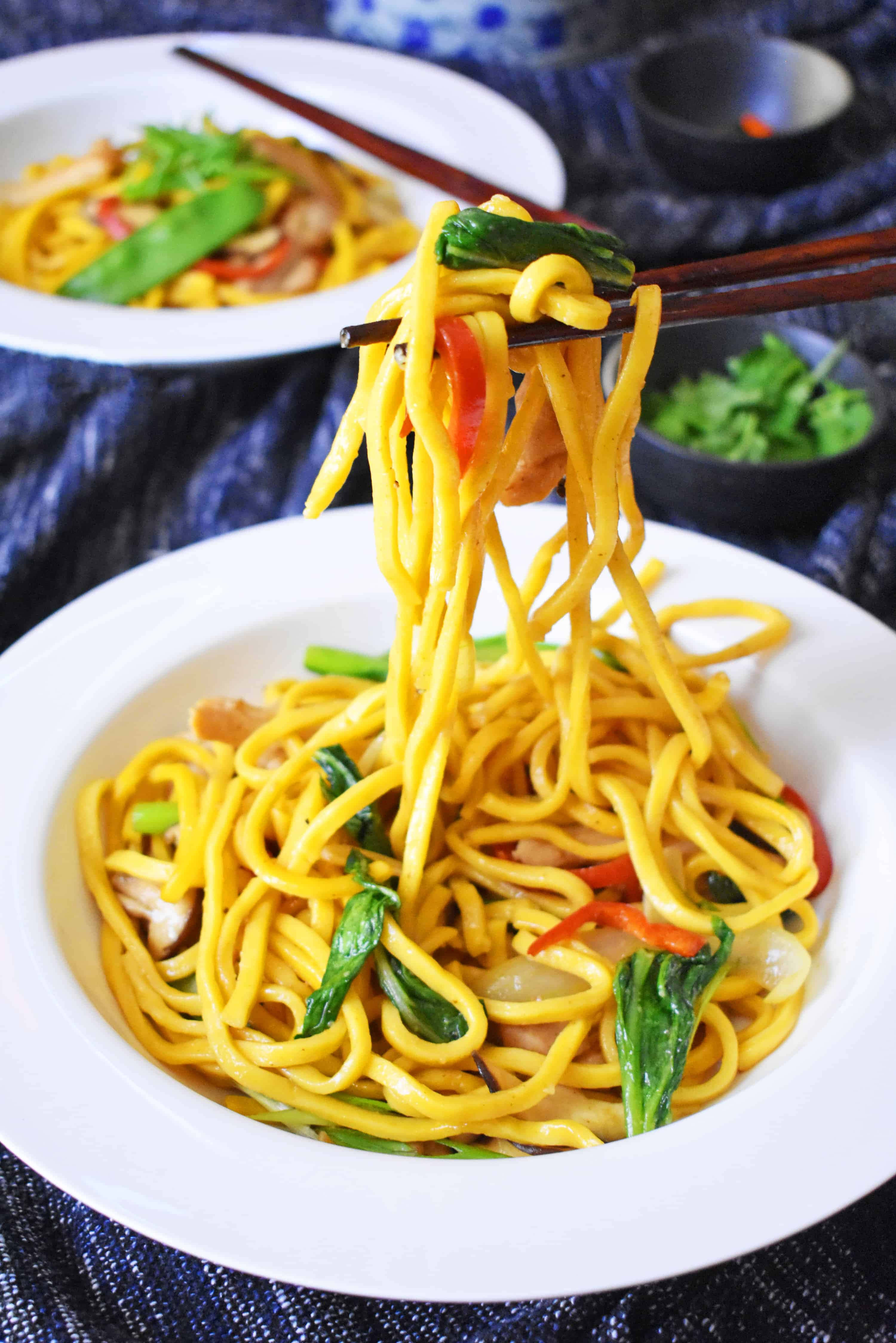 Noodles 桌布 壁紙 — 筷子從深色紋理桌上的白色碗中夾起光澤的黃色麵條，混合著色彩繽紛的蔬菜。
