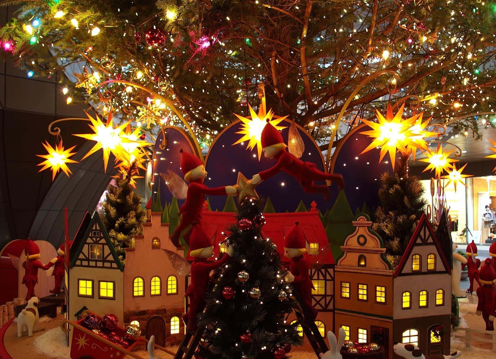 Christmas Village 桌布 壁紙 — 節日精靈在閃爍燈光和星星裝飾的天篷下裝飾發光的聖誕樹，在一個奇特的假日村莊裡。