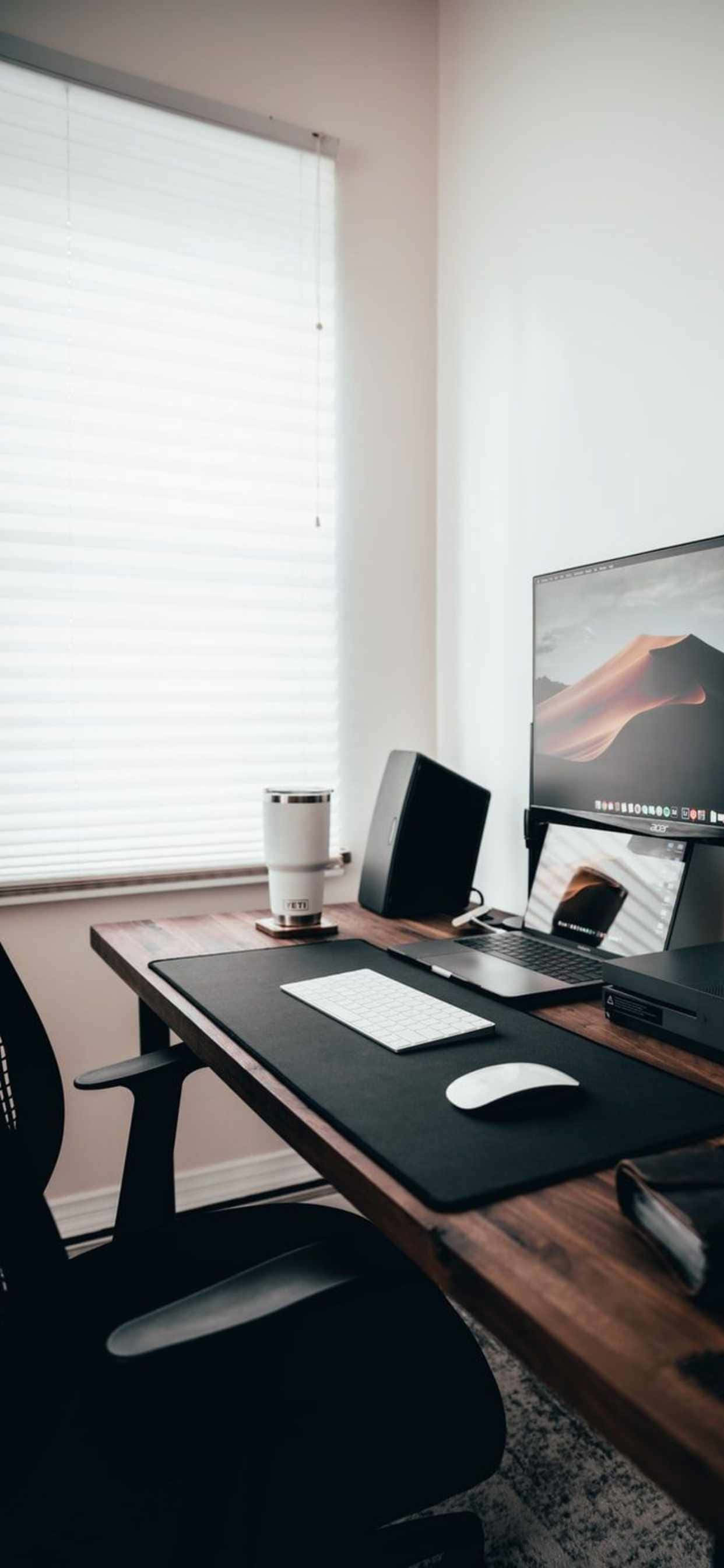 iPhone Xs Max Office 桌布 壁紙 — 簡約居家辦公室，配有木桌、黑色椅子和筆記型電腦，百葉窗透出柔和自然光。
