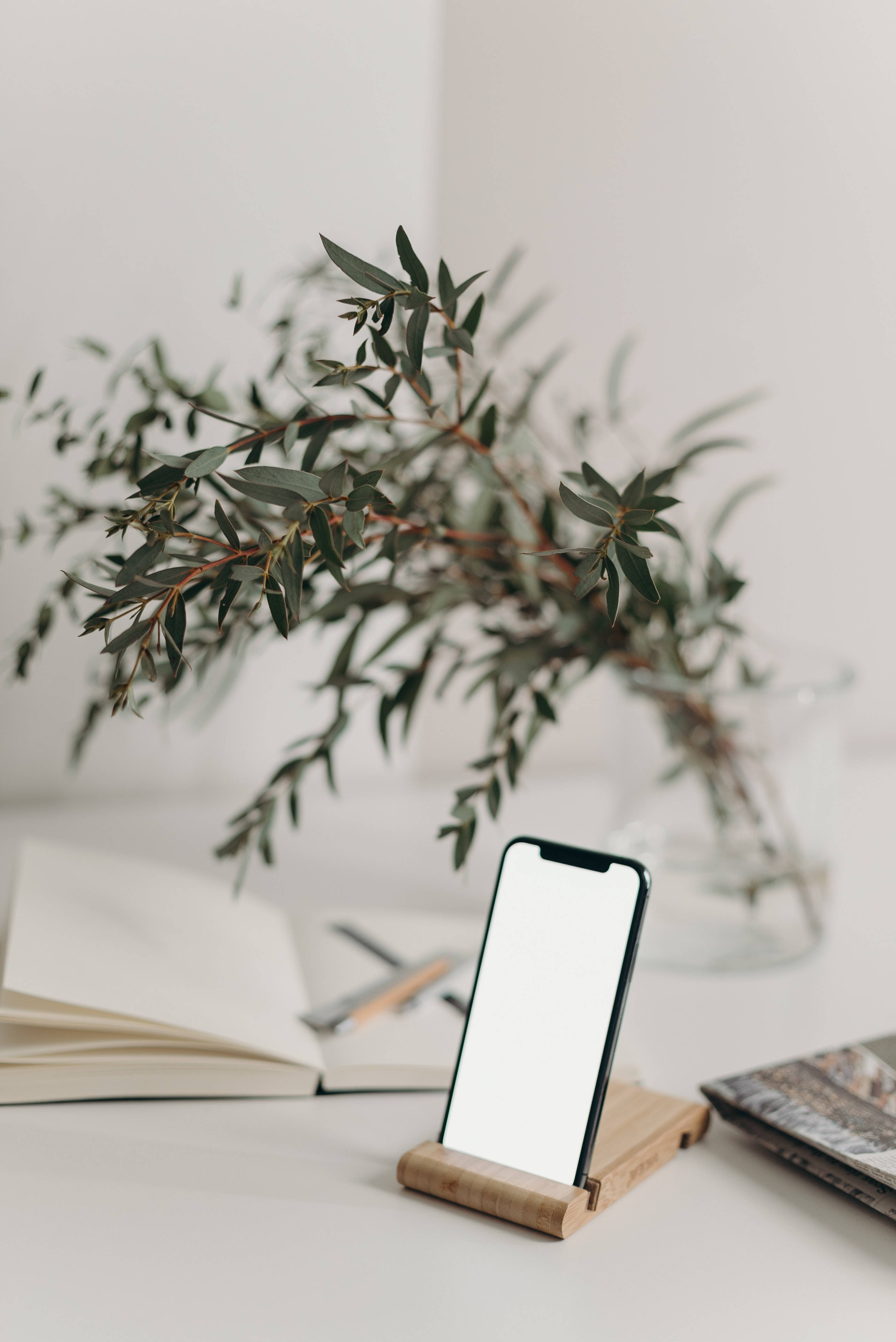 酷炫iPhone 桌布 壁紙 — 簡約書桌設置，木製支架上的智慧型手機，旁邊放著一本打開的書和玻璃花瓶中的尤加利樹枝。