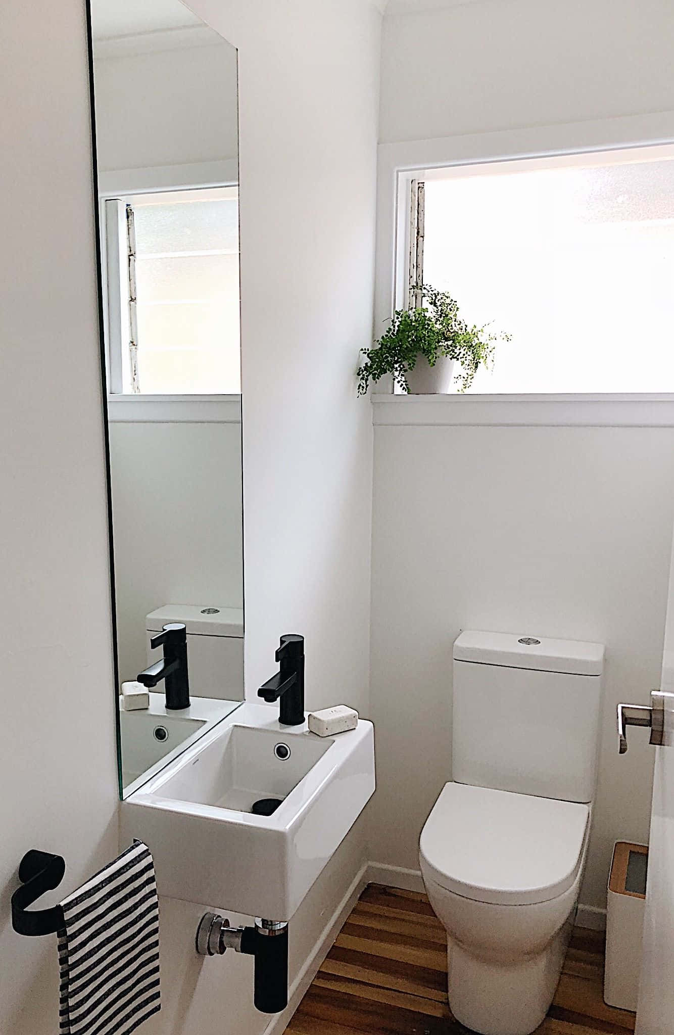 Toilet 桌布 壁紙 — 簡約浴室，配有白色衛浴設備、黑色水龍頭、木地板和窗邊的綠色植物。