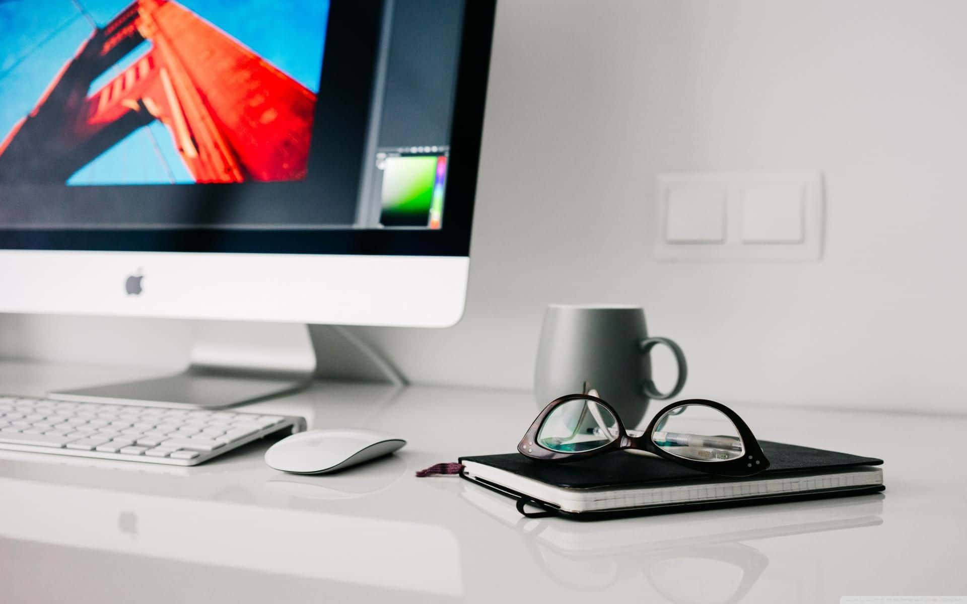 Eyeglasses 桌布 壁紙 — 簡約白色桌面設置，iMac 顯示鮮紅色橋樑壁紙，眼鏡，馬克杯和筆記本。