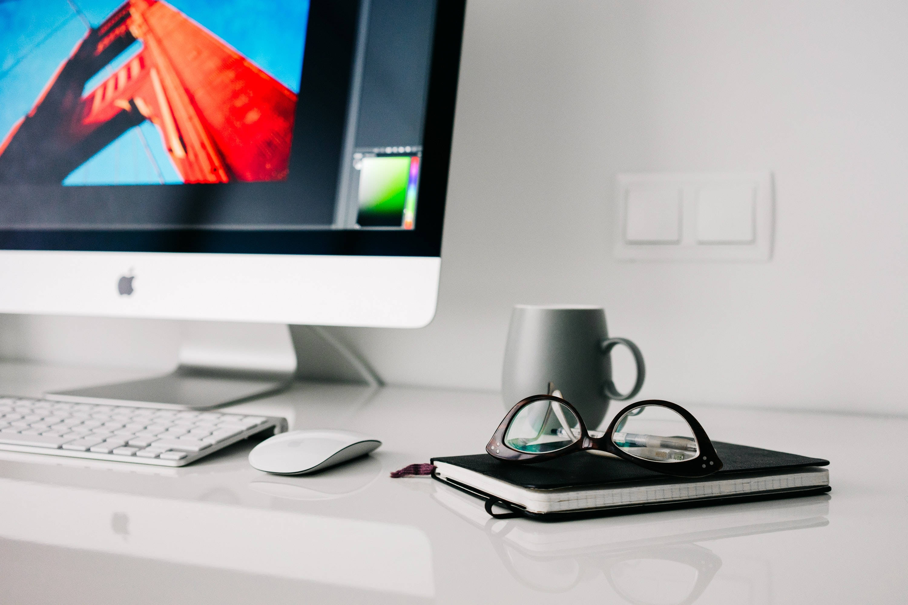 Mac 桌布 壁紙 — 簡約白色桌面設置，iMac 顯示紅色橋樑桌布，灰色馬克杯和眼鏡放在筆記本上。