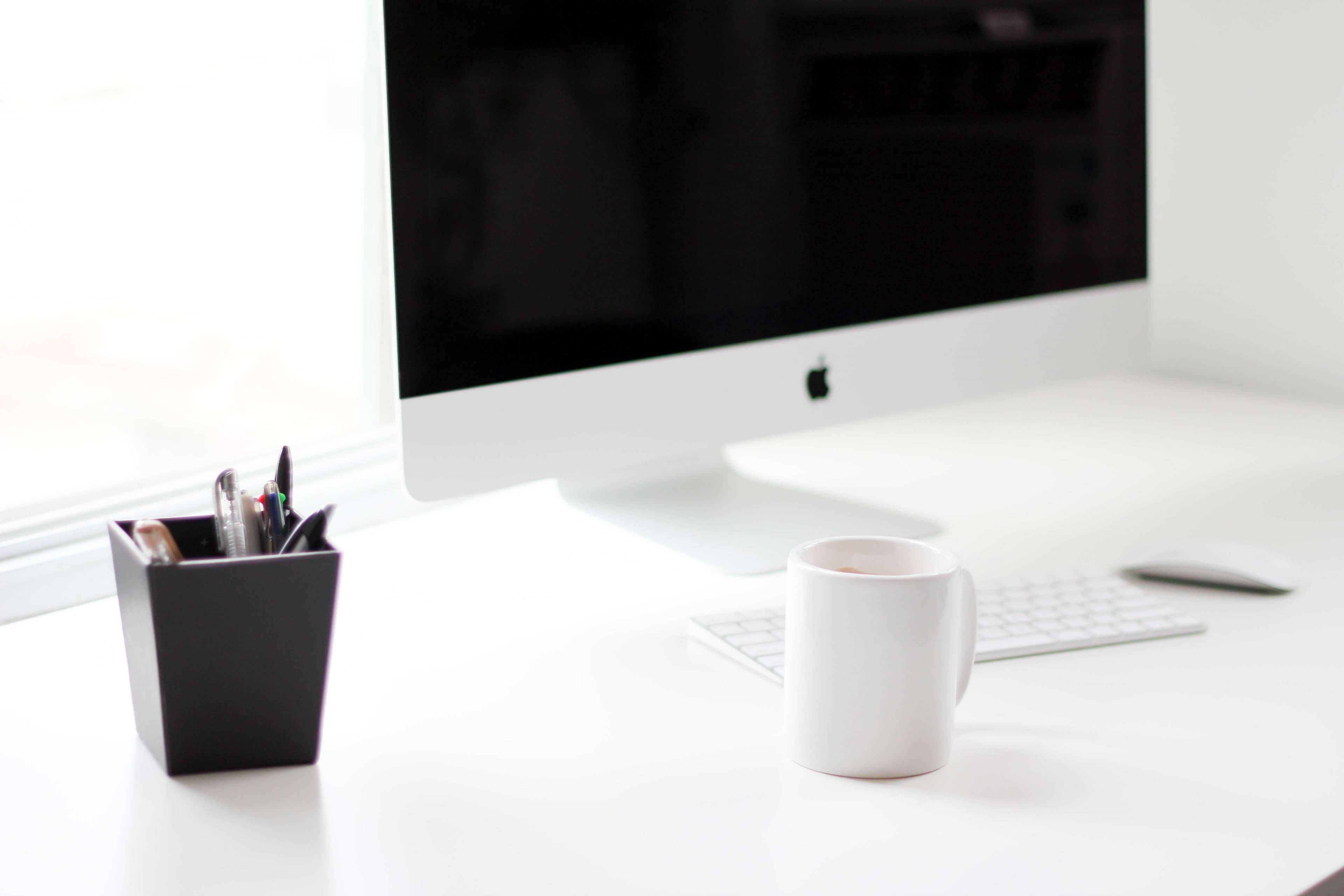 Work 桌布 壁紙 — 簡約白色桌面設置，配備 iMac 鍵盤、滑鼠、馬克杯和黑色筆筒，沐浴在柔和自然光中。
