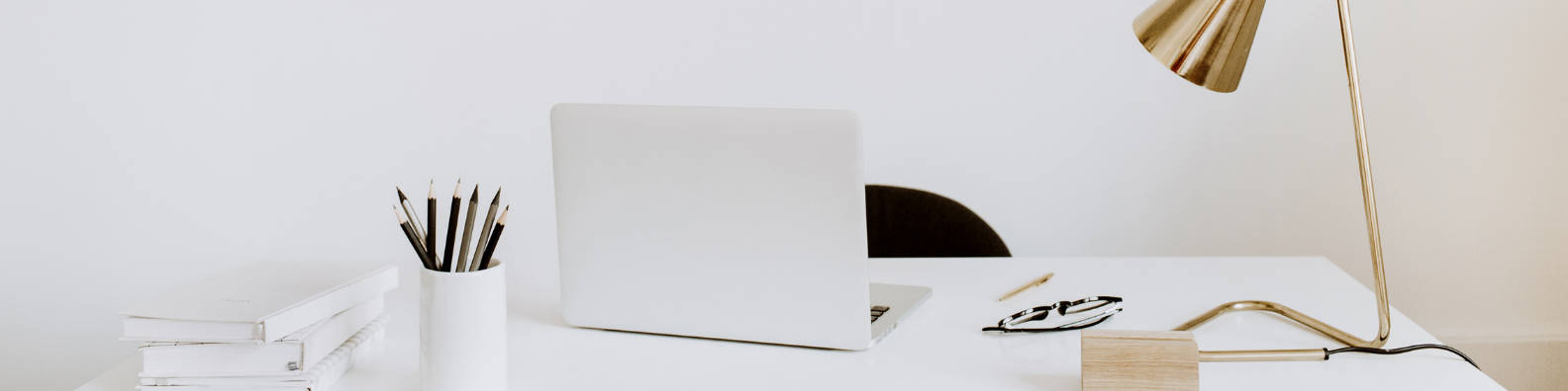 Office Desk 桌布 壁紙 — 簡約白色桌面設置，配備筆記型電腦、金色檯燈和疊放的書籍，柔和自然光線下。