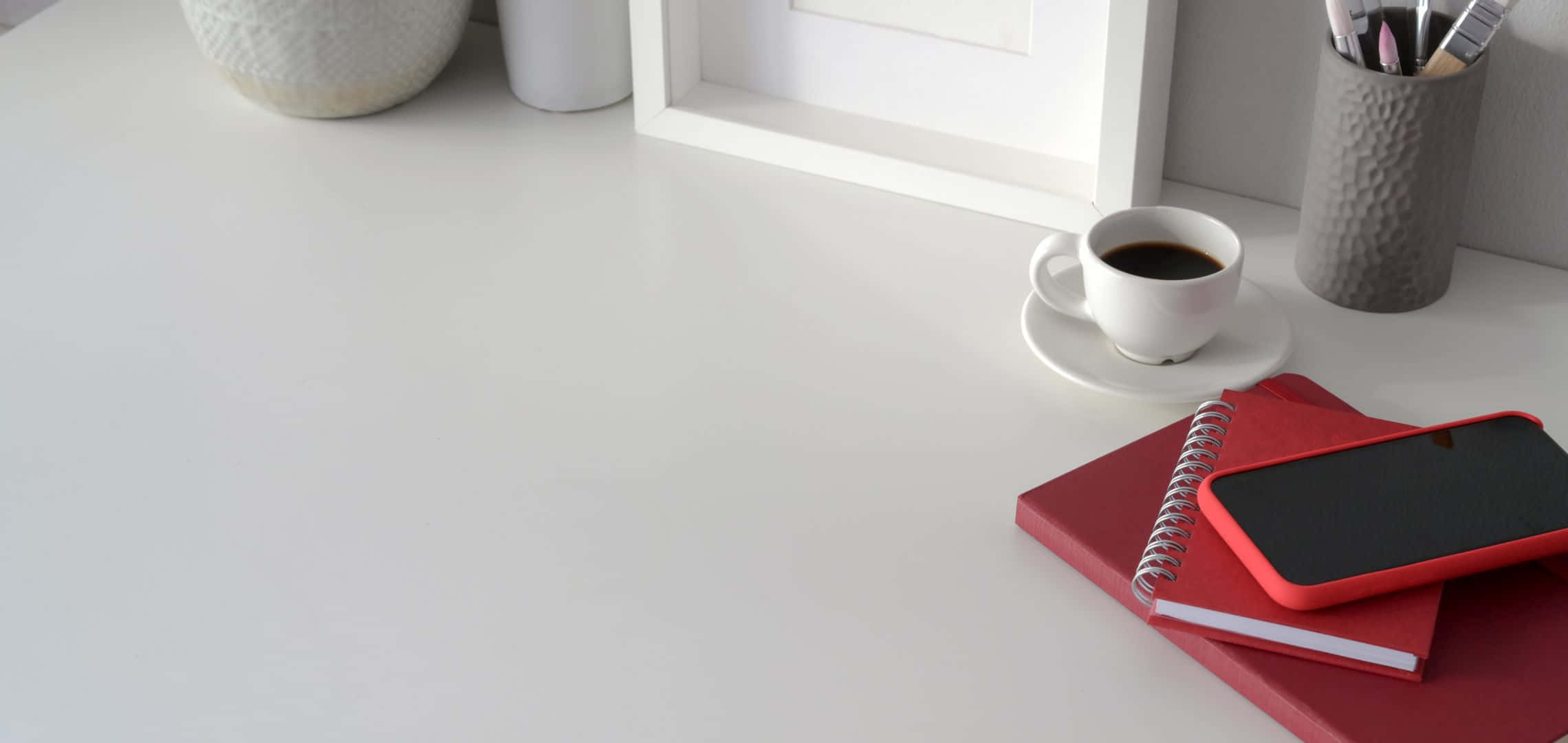 2280x1080 桌布 壁紙 — 簡約白色桌面設置，配有紅色筆記本、電話、咖啡杯和裱框藝術品，營造平靜的工作空間氛圍。