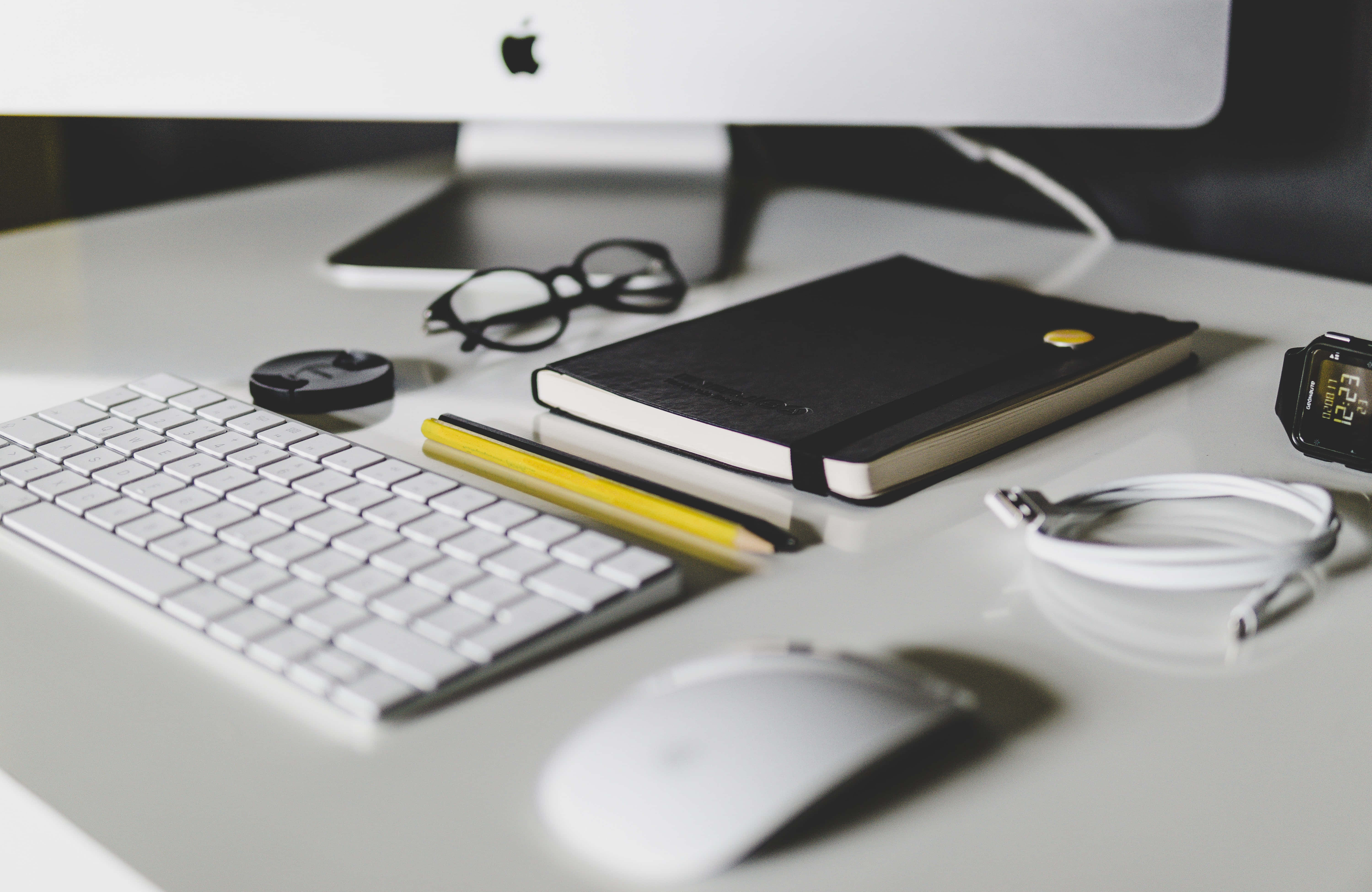 Computer Mouse 桌布 壁紙 — 簡約白色桌面設置，配有鍵盤、滑鼠、筆記本和眼鏡，在柔和日光下。