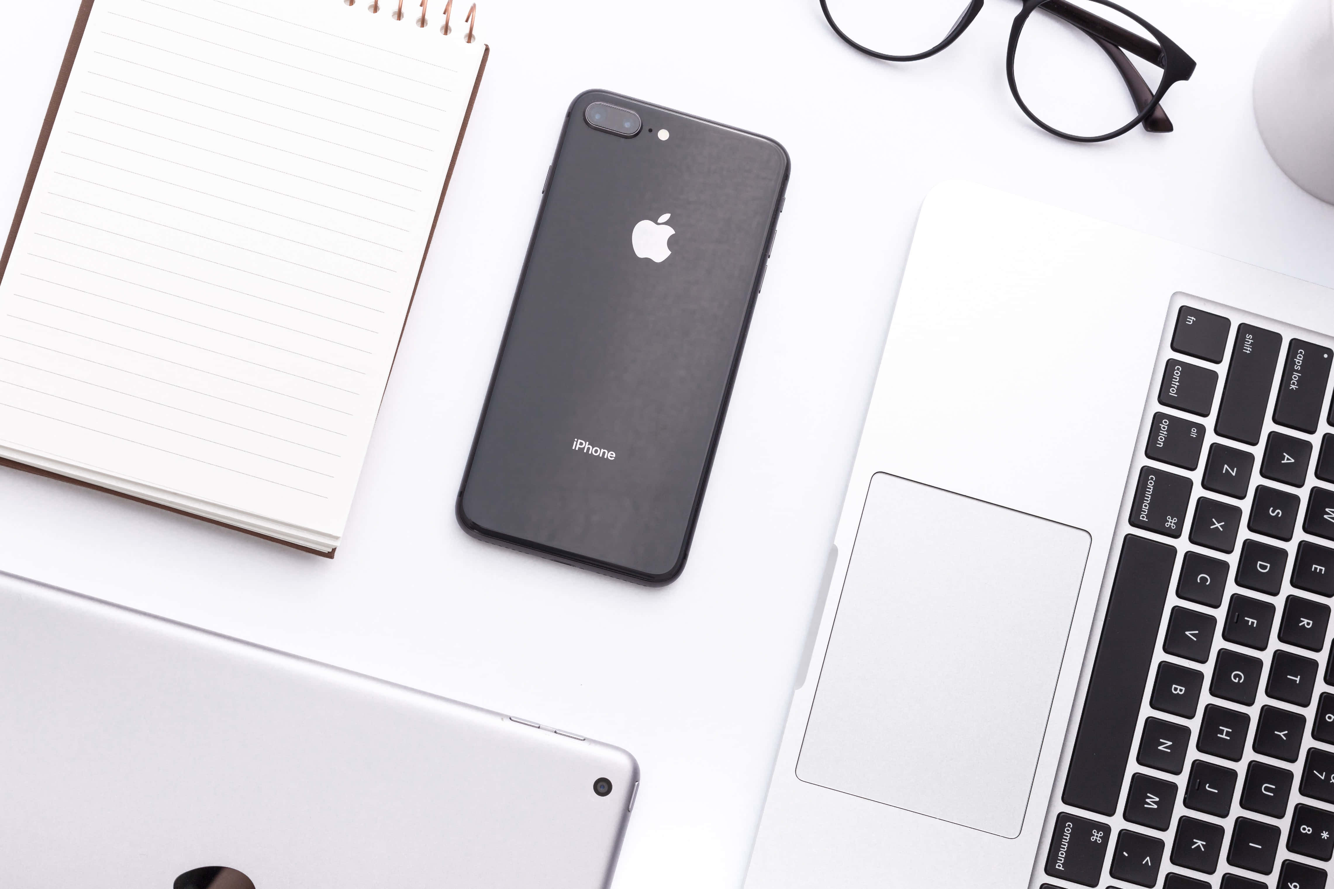 iPhone 桌布 壁紙 — 簡約白色桌面設置，配有黑色iPhone、筆記型電腦、筆記本和眼鏡，整潔現代。