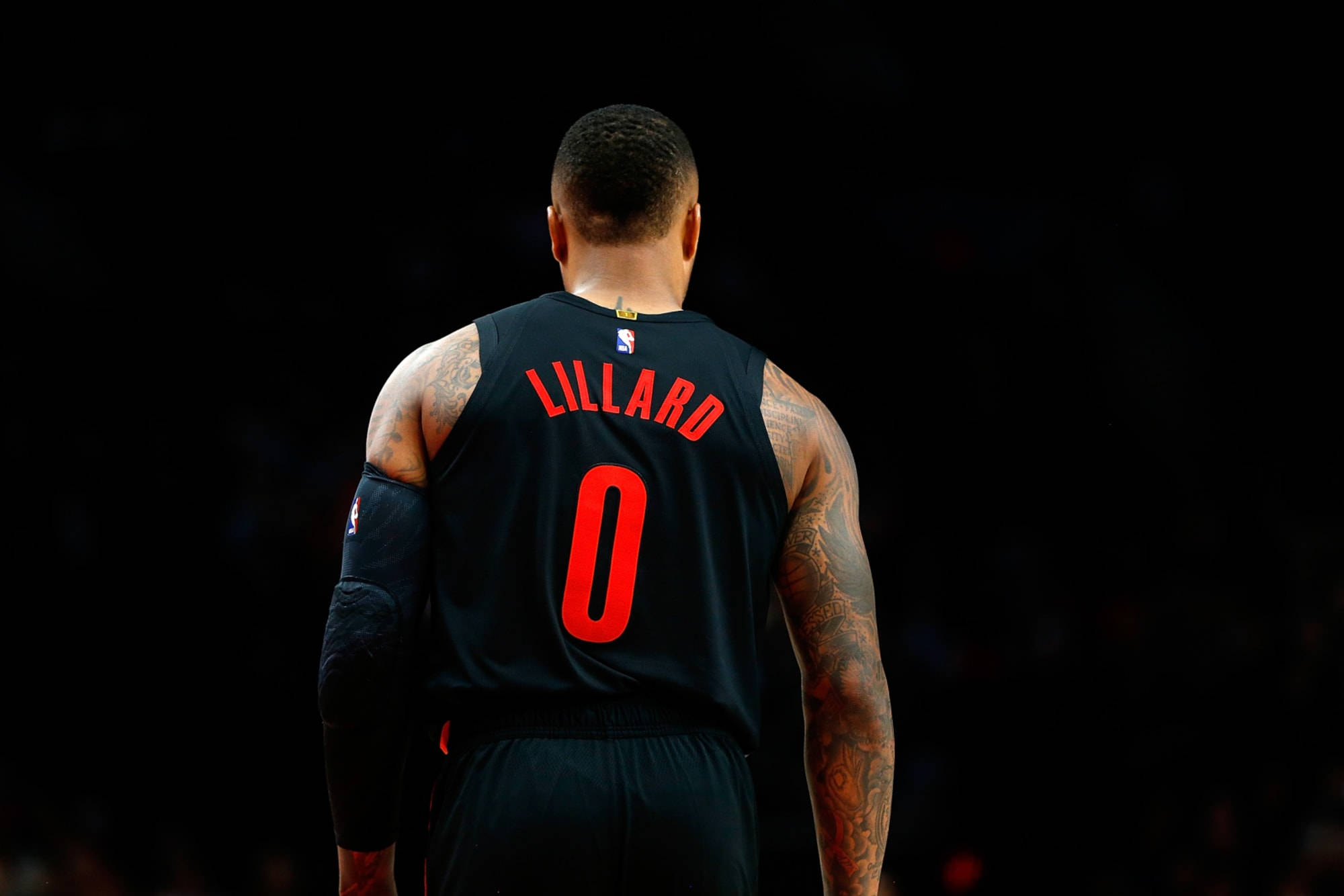 Damian 桌布 壁紙 — 籃球運動員 Damian Lillard 身穿黑色球衣，紅色零號，紋身清晰可見，背對深色背景。